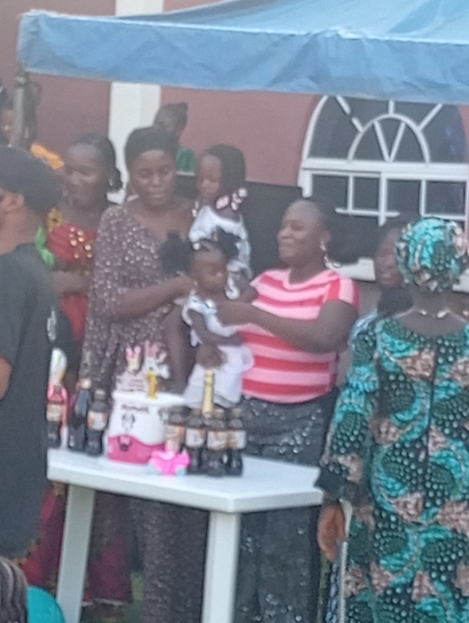 A gathering is taking place under a canopy in what appears to be an outdoor event in Rido, Nigeria. Several women are gathered around a white table. A birthday cake and several bottles of Coca-Cola are on the table. Two women are holding young children. One woman is wearing a pink and red striped shirt, while another is wearing a floral-patterned dress. A woman in a green and brown patterned dress stands to the right. A building with an arched window is visible in the background.