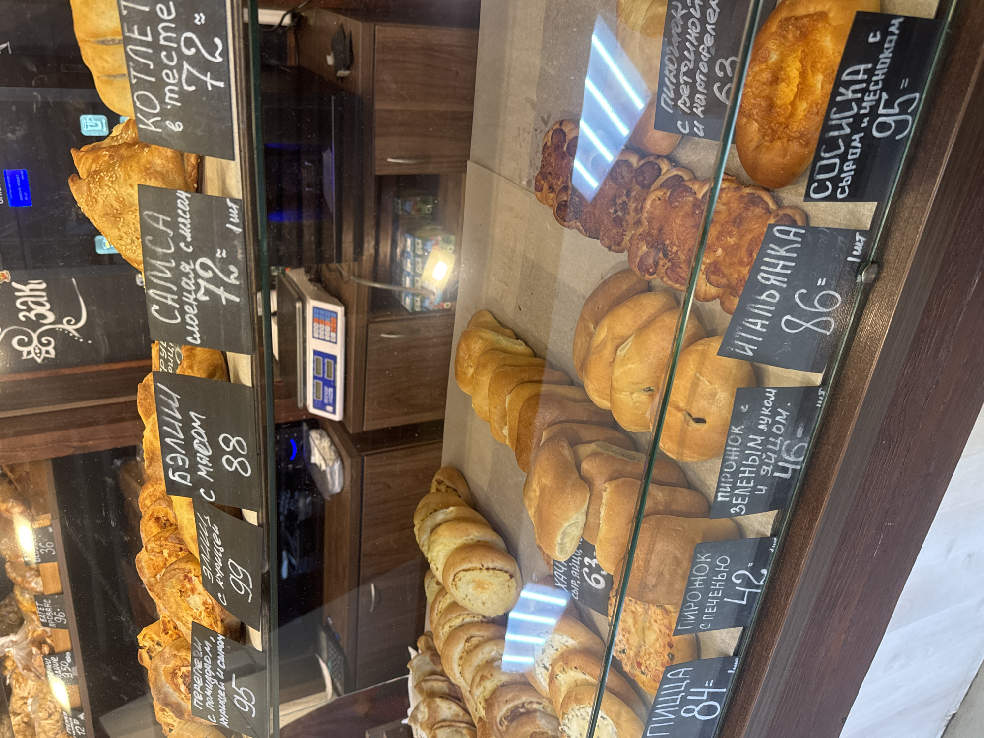 A refrigerated display case showcases a variety of baked goods with handwritten price tags, indicative of a bakery or food shop, likely located in the city of Chaikovsky, Russia. The food items range from small rolls to larger pastries filled with meat, vegetables, cheese, and sausage. Tags indicate prices in Russian rubles, next to each item. A set of scales sits on the display case.