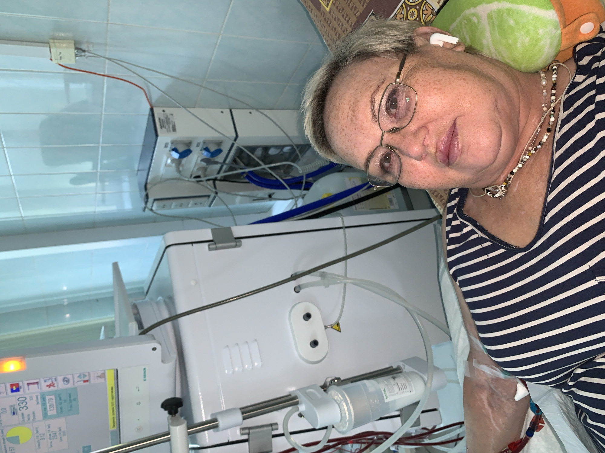 A woman lies in a hospital bed, possibly undergoing dialysis. She is wearing a striped shirt and glasses, and has earbuds in. The bed is positioned next to a dialysis machine and other medical equipment, including monitors and tubing. The room appears to be tiled, with fluorescent lighting. The image is taken from a slightly elevated angle, looking down at the woman.