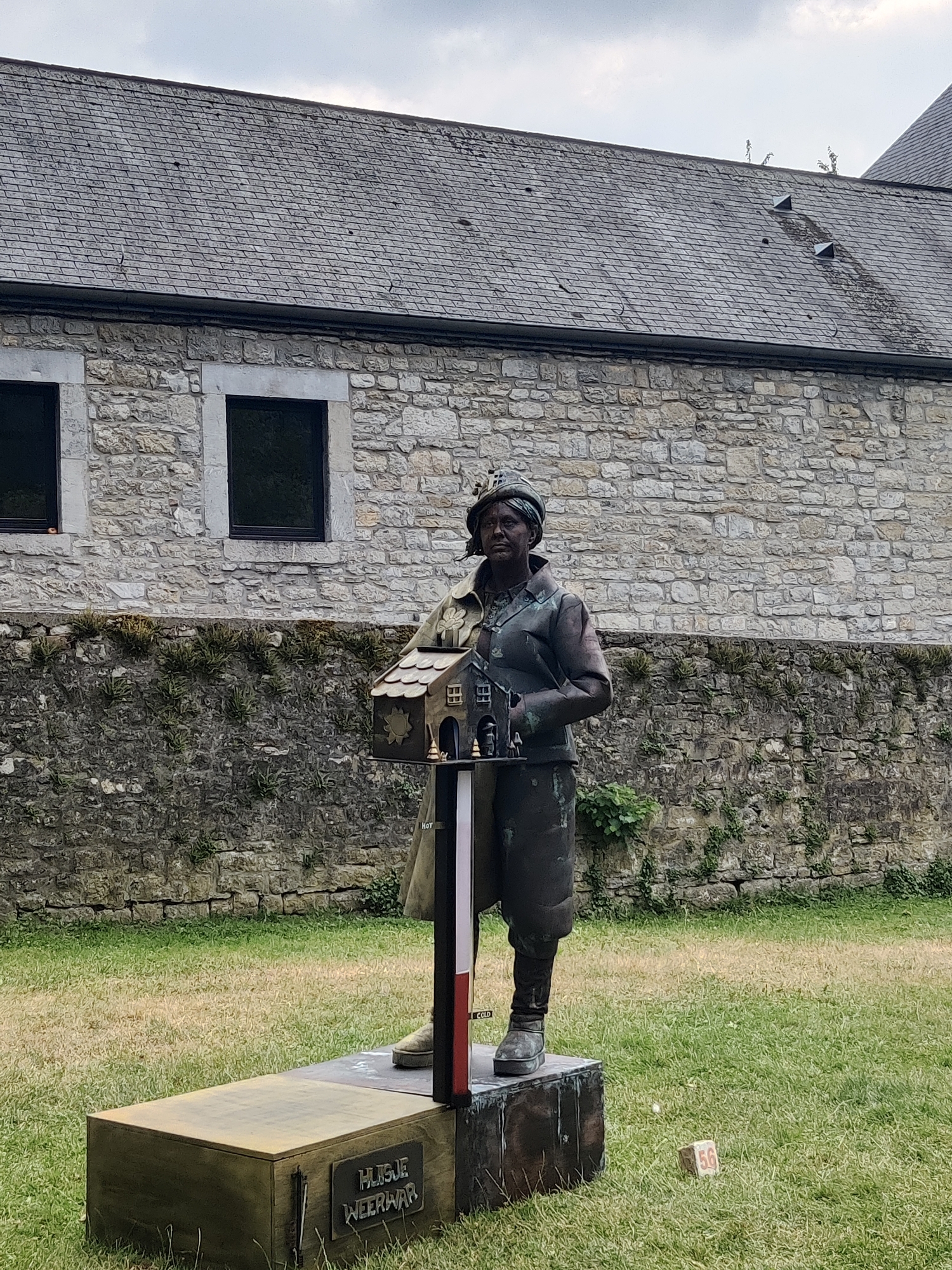 A bronze statue of a woman wearing a military-style hat and uniform stands on a pedestal in a grassy courtyard. The woman is holding a miniature house in her left arm. The statue is located in front of a stone building with a grey tiled roof. A sign on the pedestal reads "HUGUE WEERWAR." The building appears to be a historic structure, possibly a castle or old manor, with visible age and weathering on the stone and roof. The courtyard is covered in grass, with some patches of dry, yellowed grass interspersed with greener areas. The sky is overcast, suggesting a cloudy day. A small marker with the number "56" is visible on the grass to the right of the statue. The overall scene evokes a historical or memorial setting. This statue is located in Marche-en-Famenne, Belgium, specifically in the Parc de la Poste, and it commemorates the role of women during the war.