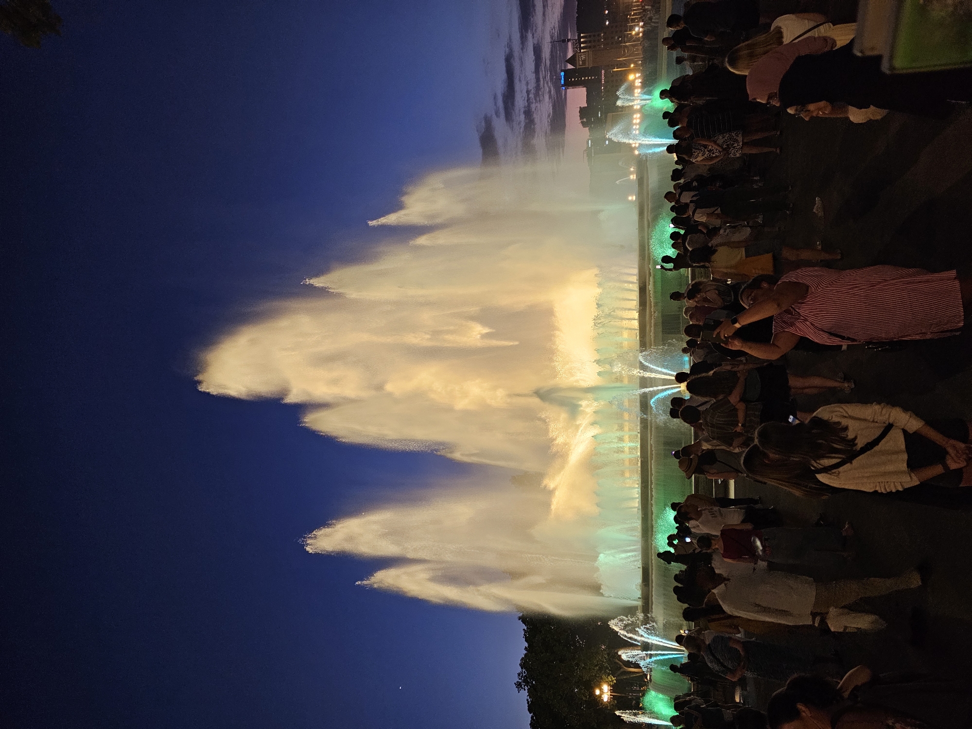 The image captures a night scene in Barcelona, Spain, likely at the Magic Fountain of Montjuïc, showcasing a spectacular water and light show. A large crowd of people is gathered, facing the fountain, with many holding up their phones to take pictures or videos. The fountain itself is the central subject, with massive plumes of water illuminated in warm golden and cool green lights, creating a dramatic and vibrant display against the dark blue twilight sky.
The foreground is filled with a diverse group of individuals, appearing to be tourists or locals enjoying the performance. They are dressed in casual evening wear. Some are standing, while others are seated or walking. Their attention is directed towards the fountain. The overall atmosphere is one of shared enjoyment and wonder.
In the background, the outlines of buildings are visible on the horizon, suggesting an urban setting. The sky transitions from a deep blue overhead to lighter hues near the horizon, indicating the end of dusk. Trees and lampposts are also present, contributing to the public space ambiance. The water jets are caught in motion, appearing as soft, luminous shapes, and the lighting creates a sense of depth and dynamism.