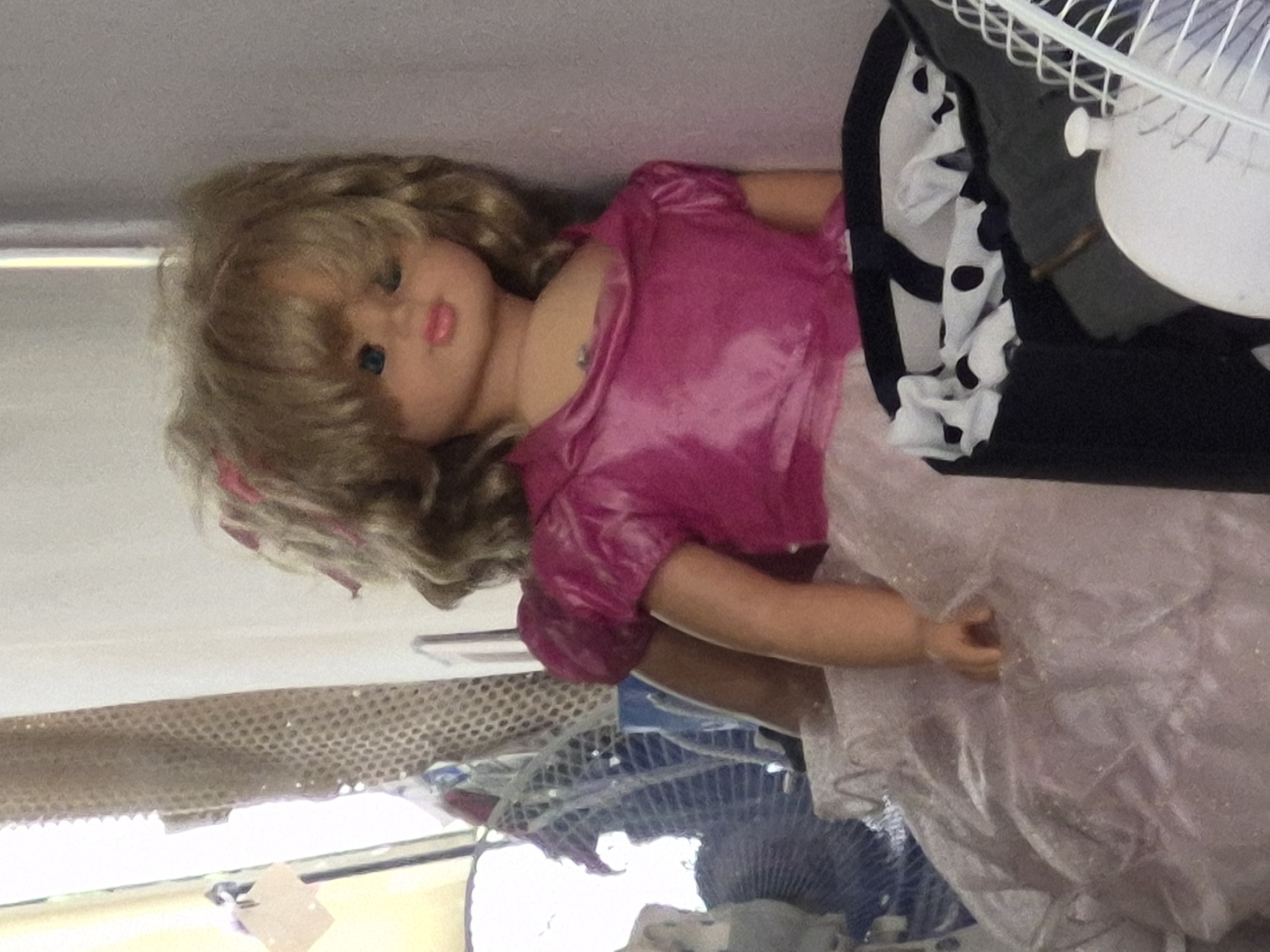 The photograph displays a blonde doll lying horizontally, positioned with its head towards the left and body extending to the right. The doll has wavy blonde hair, blue eyes, and pink lips. It is dressed in a shiny, short-sleeved fuchsia or bright pink top, and the lower part of its outfit consists of a light, possibly sheer and crinkled fabric, resembling a skirt or dress. One of the doll's arms is slightly bent, with its hand visible among the fabric.
To the right of the doll's body, a white electric fan with a visible safety grille is partially seen. Behind the fan, there's a folded piece of black fabric adorned with white polka dots or a similar pattern. The background on the left and lower sections includes parts of other grates or fan structures, suggesting a cluttered indoor environment. The upper portion of the image features a plain, light-colored surface, likely a wall or ceiling.
The scene is a static still life, with no discernible activity or event taking place. The lighting suggests an indoor setting, likely during the daytime. There is no visible text in the image. This arrangement of items is observed in what is understood to be a location in Yerevan, Armenia.