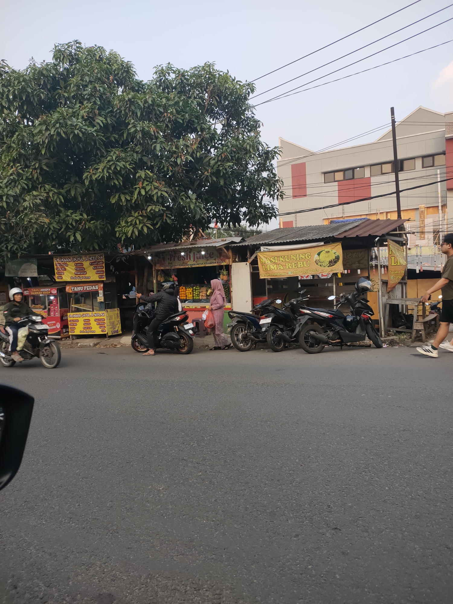 The image captures a street scene in Cimahi, Indonesia, likely in the afternoon or early evening due to the lighting. In the foreground, a dark asphalt road is visible, with the reflection of a car's side mirror on the left. Along the right side of the road is a row of small roadside stalls, one of which displays a banner for "Fresh Juice" with text in Indonesian. Oranges and other fruits are visible in the stall. Several motorcycles are parked or in motion near the stalls.
On the left side of the street, a person wearing a helmet rides a motorcycle. Another person wearing a helmet is on a stationary motorcycle, pointing. A person in a pink headscarf and dress is walking near the fruit stall, carrying a plastic bag. To the right, a man wearing a dark green t-shirt and shorts walks away from the stalls.
Behind the stalls, a large green tree with dense foliage dominates the left side of the frame. Further in the background, a modern building with a light gray facade and red rectangular accents is visible, with utility wires crisscrossing in front of it. The sky is overcast, with a pale blue hue. The overall impression is of a busy, functional street-level commercial area.