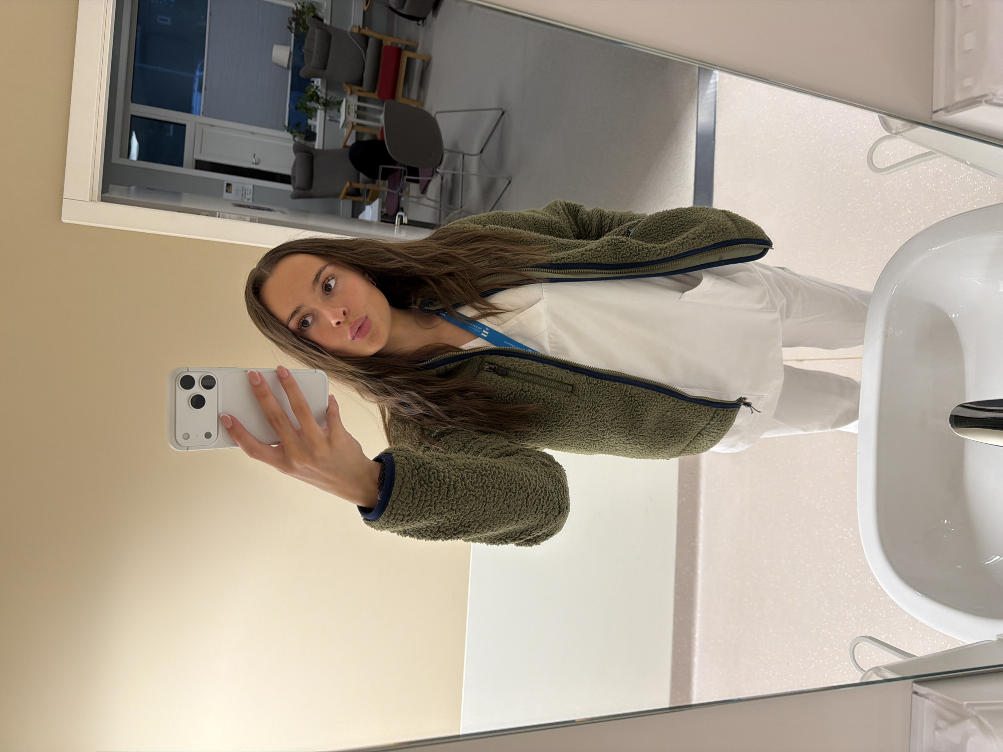 A young woman with long brown hair takes a mirror selfie in what appears to be a restroom or changing area within a medical facility. She is wearing a white scrub top and white scrub pants, with an olive green fleece jacket zipped over the scrub top. A blue lanyard with an identification card is visible around her neck. She is holding a white smartphone in her right hand, angled to capture her reflection. Her left hand is tucked into the pocket of her fleece jacket. The background reflected in the mirror shows a room with several chairs, a table, and large windows. The floor is a speckled light color, and the walls are painted a neutral beige. A white sink is visible in the foreground of the mirror. The overall impression is of a casual, everyday moment captured in a professional setting. It is not possible to determine the location in the city of Harstad, Norway from the visual information provided.
