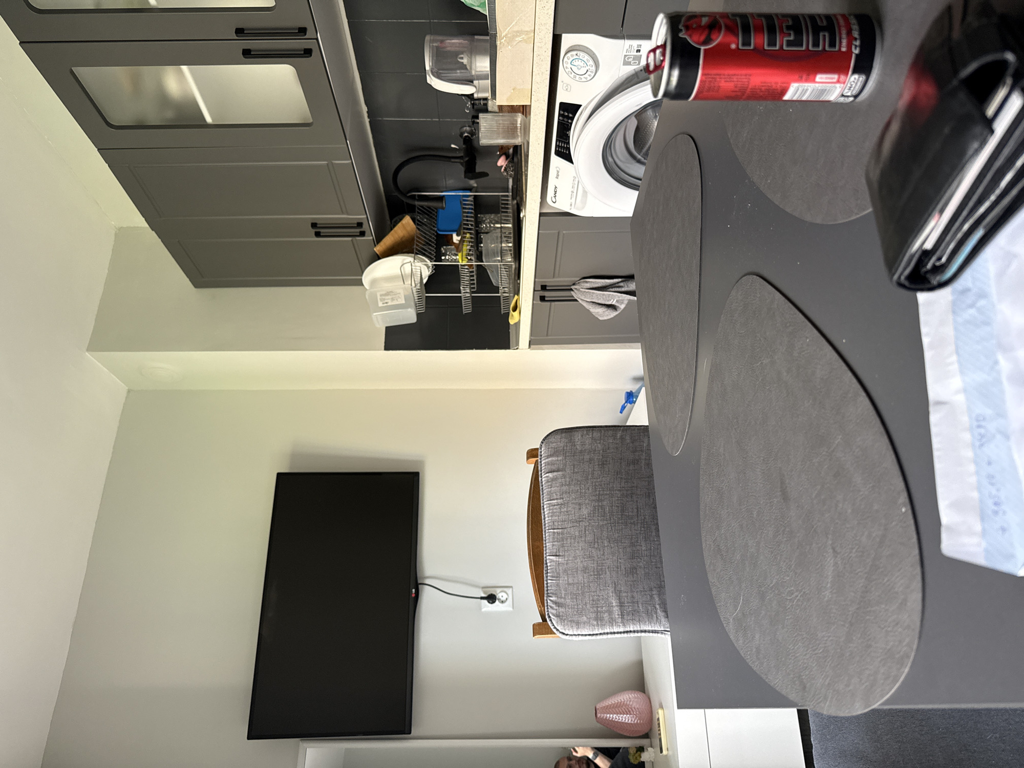 The image shows an interior scene, likely a kitchen and dining area. The kitchen area features gray cabinets, a sink with a dish rack, and a white washing machine. On the dining table, there are round placemats, a can of "Hell" energy drink, and a wallet. In the background, there is a gray wall with a mounted television and a white shelf. A gray chair is placed near the table. A pink vase is placed on the white shelf. The environment appears to be a modern apartment or house. There are no people visible in the image, although the presence of personal items such as the wallet and energy drink suggest someone lives there.
There is insufficient information to establish that the image comes from Stara Zagora, Bulgaria.