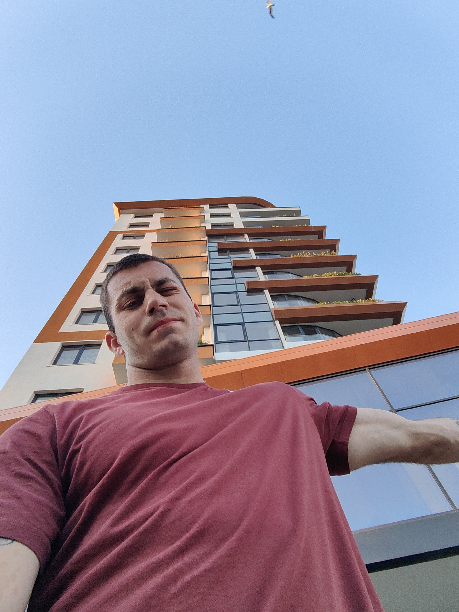 A low-angle, wide-shot selfie captures a man in the foreground, looking upwards with a slightly grimaced expression. He is wearing a maroon t-shirt. Behind him, a modern, tall building with a distinctive orange and white facade dominates the background. The building features multiple balconies with plants and large glass windows. The sky above is clear and blue, with a single bird visible in flight near the top center of the frame. The image appears to have been taken outdoors during daylight hours, possibly in the city of Varna, Bulgaria, given the architectural style and the clear sky.