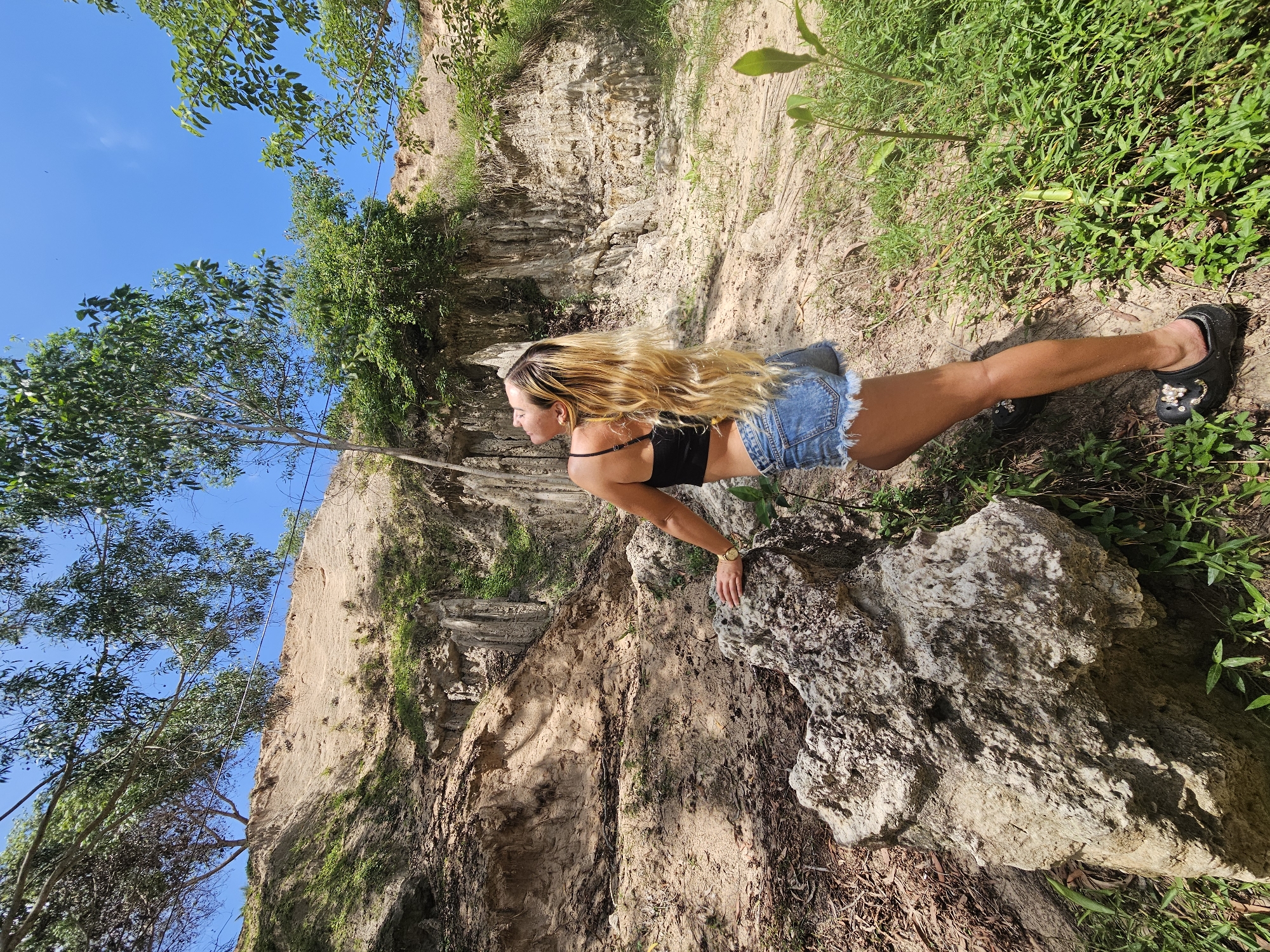 This image captures a person engaged in a push-up on a rocky, uneven terrain. The subject, a woman with long, blonde hair, is wearing a black sports bra and denim shorts. Her body is positioned in a push-up stance, with her hands resting on a large rock in the foreground. The background reveals a steep, eroded cliff face with patches of vegetation and dry soil. Lush green plants and trees frame the scene, with a bright blue sky visible in the upper left portion of the image. The sunlight suggests it is daytime, likely with clear weather, given the vibrant blue sky and strong shadows cast by the rocks and vegetation. The overall impression is one of an outdoor activity in a natural, possibly remote, environment, with the subject displaying a sense of effort and focus. The location context provided indicates this scene is in Mũi Né, Vietnam, a region known for its distinctive sand dunes and coastal landscapes. There is no visible text in the image.