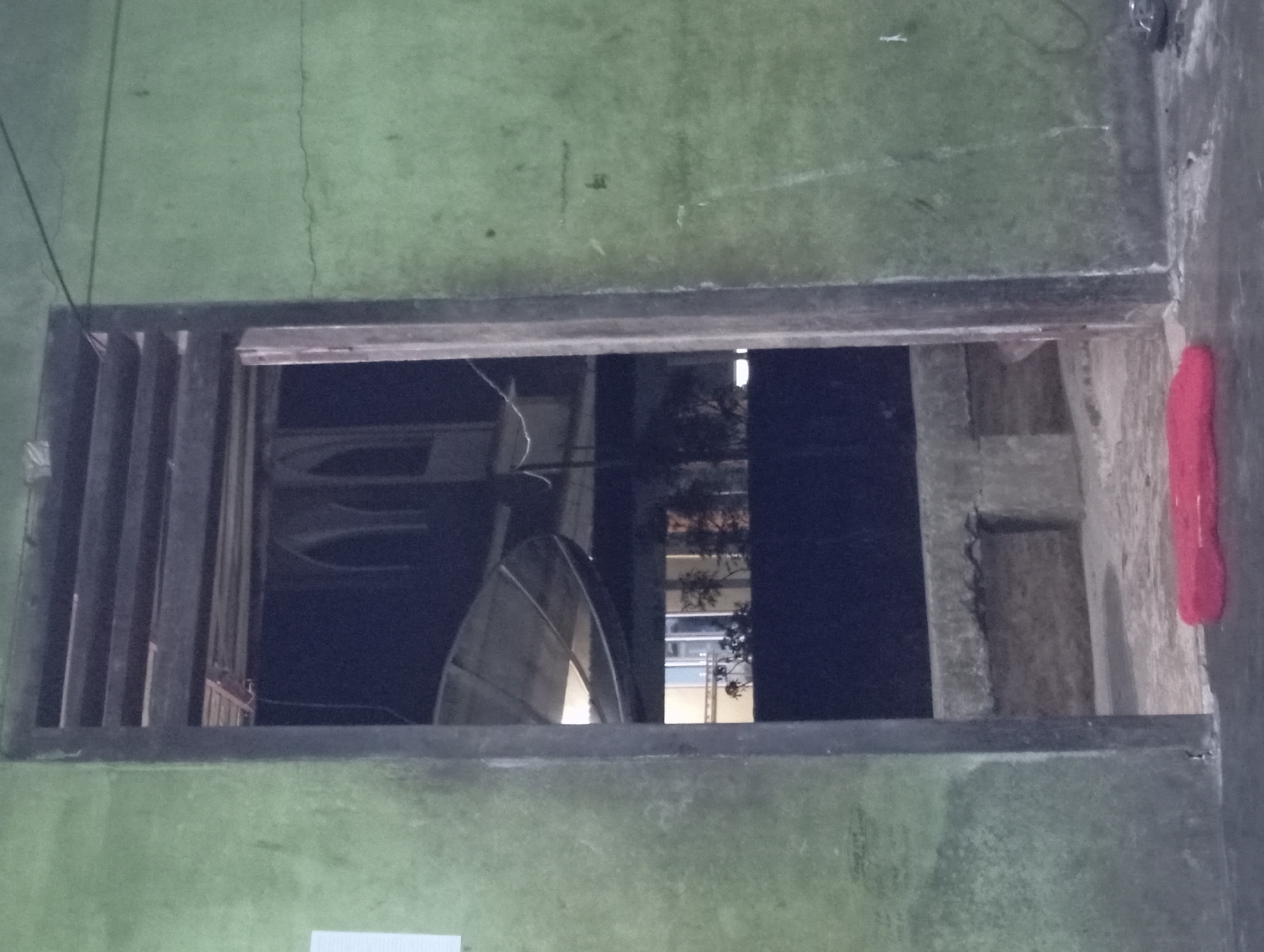 The image depicts an exterior view of a building in Rejosari, Indonesia. A doorway with wooden slats above it is visible, leading to an interior space. Through the doorway, architectural elements, possibly arches, and a satellite dish are discernible. The exterior walls of the building are painted green and show signs of wear and tear, including cracks. In the foreground, a red mat or rug lies on the ground. The scene appears to be captured at night or in low light conditions.
