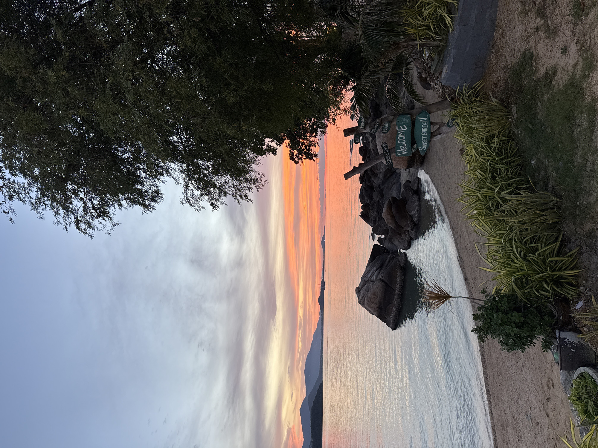 The image depicts a serene sunset scene on a beach in Baan Plai Laem, Thailand. The sky is ablaze with vibrant hues of orange, pink, and purple, reflecting on the calm ocean. In the foreground, a sandy beach is bordered by lush green vegetation and a large tree casting a shadow. Several rocks are scattered along the shoreline, and a signpost with directions to "Sunset Garden," "Koh Phangan," "Phuket," and "Hua Hin" stands prominently. The overall atmosphere is peaceful and tranquil, capturing the beauty of a tropical evening. There are no people or animals visible in the image.