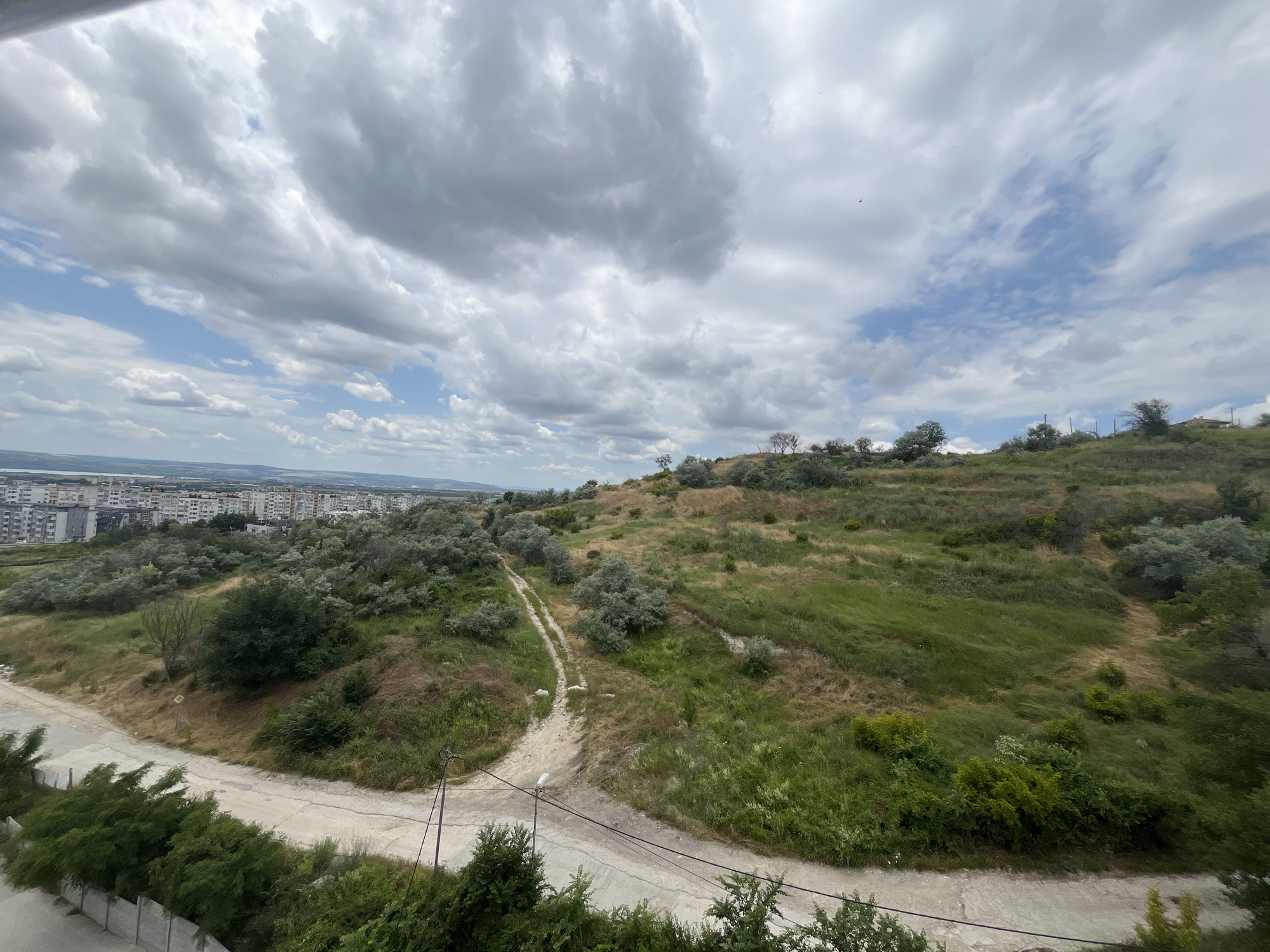This is a wide-angle, high-angle shot of a semi-urban landscape on a cloudy day, likely taken from a building in Varna, Bulgaria. The foreground features a concrete pathway and a low concrete wall, with dense green foliage obscuring the very bottom of the frame. A dirt path winds uphill into a grassy, undulating terrain dotted with scrubby bushes and a few sparse trees. In the mid-ground, the landscape transitions to a more urban environment with a cluster of multi-story apartment buildings visible to the left. The background is a hazy expanse of land and sky. The sky is filled with dramatic, heavy gray clouds interspersed with patches of blue. The overall impression is of a transition zone between a somewhat neglected natural area and a residential urban district.