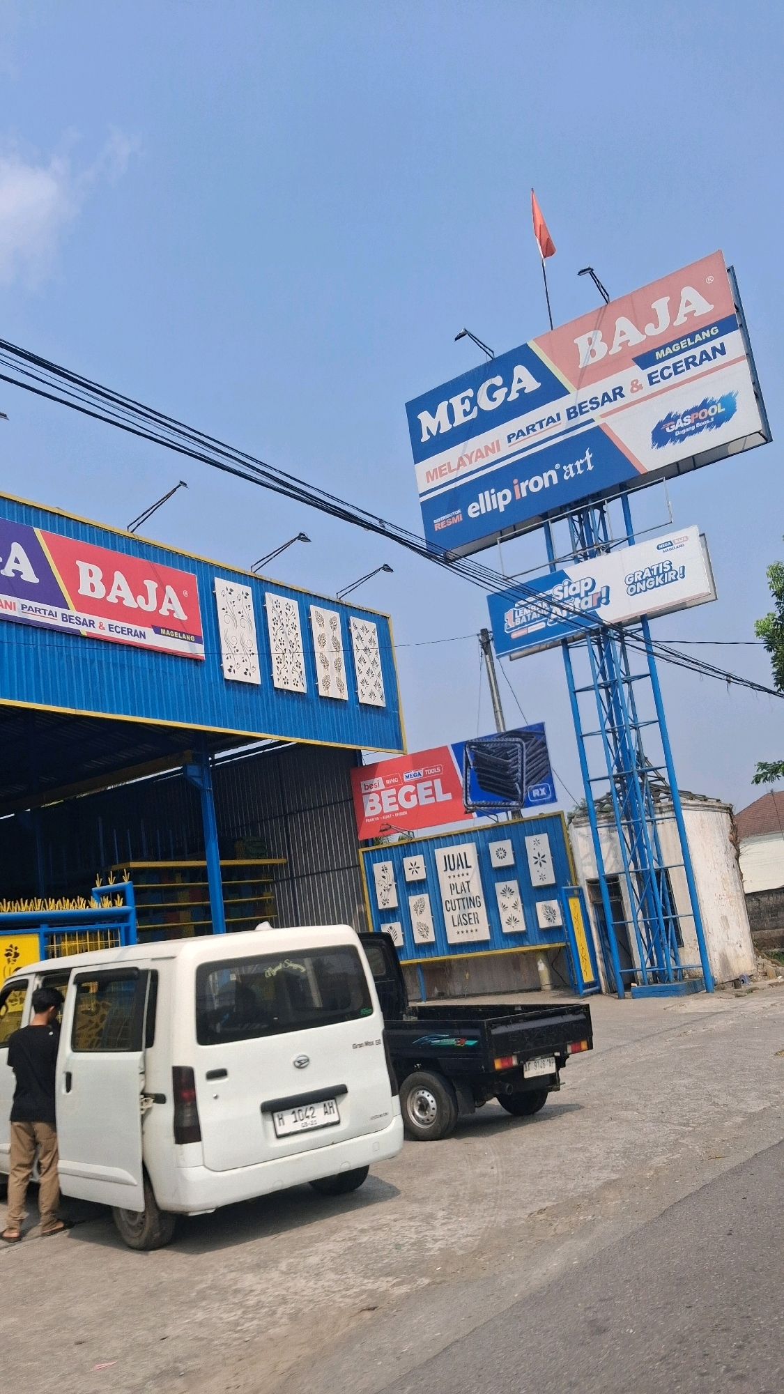 The image shows a street scene in Pabelan, Indonesia, featuring a storefront and billboards. A white minivan with its doors open is parked on the right side of the road, and a person is standing next to it. Behind the minivan, a black pickup truck is also parked. Several billboards are visible, advertising businesses such as "MEGA BAJA" and "BEGEL." The storefront appears to be a commercial establishment selling construction materials, with items like metal grating and decorative panels displayed. The sky is blue with some clouds, and power lines are strung overhead. The overall environment suggests a commercial area in a developing urban setting.