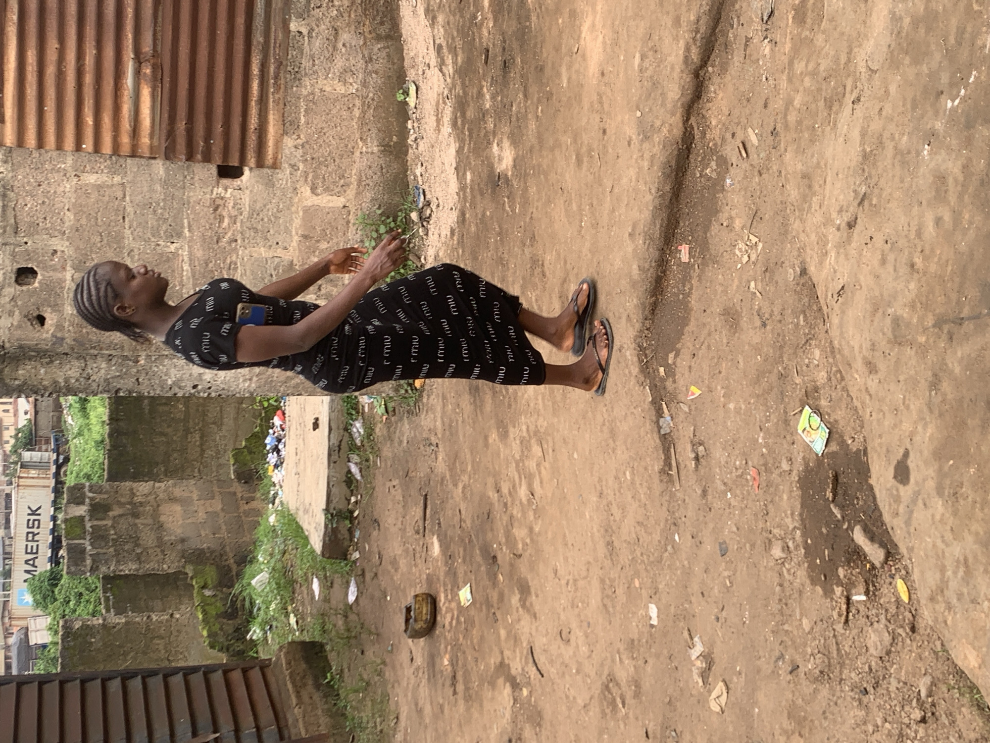 A young Black woman with braided hair walks to the right, wearing a black dress with white lettering and black flip-flops. She has a phone clipped to her waistband. She is walking on a dirt path in a sparsely populated urban area in Shagamu, Nigeria. The path is littered with debris, including discarded wrappers and a broken yellow object. In the background, there are unfinished brick buildings and a corrugated metal fence. A large white shipping container with "MAERSK" printed on it is visible in the distance, along with green vegetation.