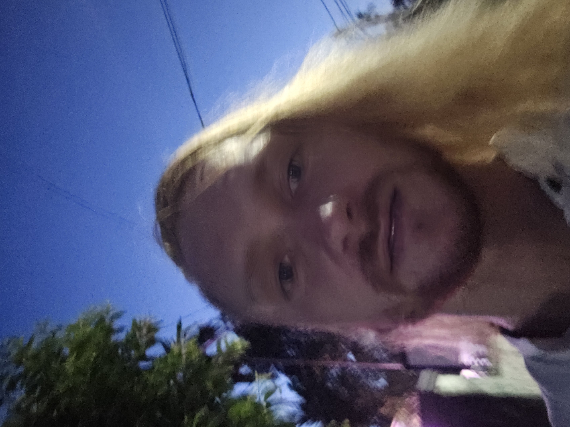 A selfie-style, slightly blurred, outdoor, waist-up photograph shows a person with long, blonde hair, light skin, and a beard. The person is looking directly at the camera, with their mouth slightly open, as if speaking or about to speak. Their hair falls over their right shoulder. The background is a darkening blue sky with some trees on the left side, and what appears to be a street or building with colored lights on the far left. The lighting suggests it is dusk or early evening. The scene does not provide any specific landmarks or visual cues to identify the location as Đà Nẵng, Vietnam.