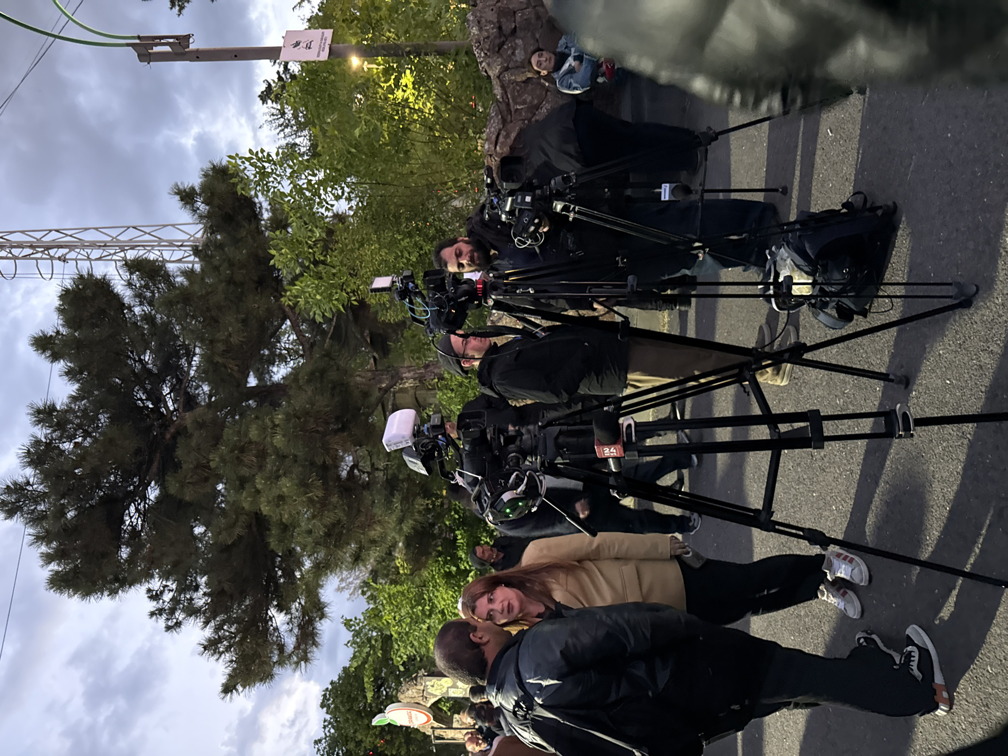 The image shows a street scene in Yerevan, Armenia. Several professional video cameras on tripods are positioned in a row, suggesting a media event or news coverage. People, including camera operators and individuals possibly being interviewed, are present. The background includes trees, a lamppost, and an overcast sky. The ground is paved.