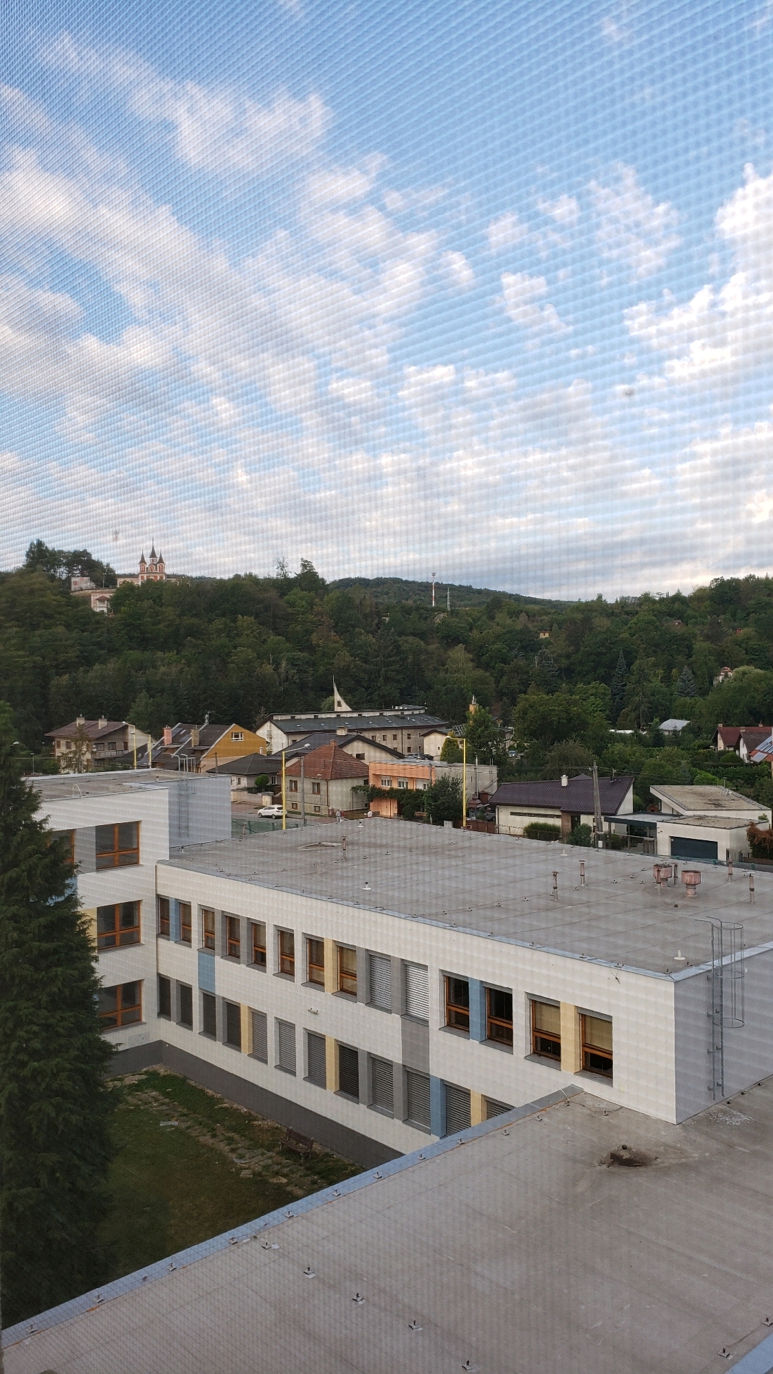 The image shows a view of a town or city from an elevated perspective, likely a window with a screen. In the foreground, there is the roof of a modern building with a gray, flat surface. To the left, part of a larger building is visible, characterized by a white facade and multiple rows of windows with brown frames. Some of these windows have colored panels (blue and yellow) between them. Adjacent to this building on the left is a tall, dark green evergreen tree.
Beyond the foreground buildings, a neighborhood of houses with red-tiled roofs and yellow or cream-colored walls is visible. Further in the distance, a densely wooded hill rises, dotted with more houses. At the very top of the hill, a prominent building with white walls and red roofs, possibly a church or a historical structure, stands out. The sky above is blue with scattered white clouds.
The scene appears to be a calm, daytime view of a residential area situated within a hilly landscape. The architecture suggests a European setting. Based on the prominent church on the hill and the overall landscape, this scene is likely located in the vicinity of the Church of St. Nicholas or the Zemplin Museum, which are notable landmarks in the city of Prešov, Slovakia, known for their elevated positions overlooking the city and surrounding hills.