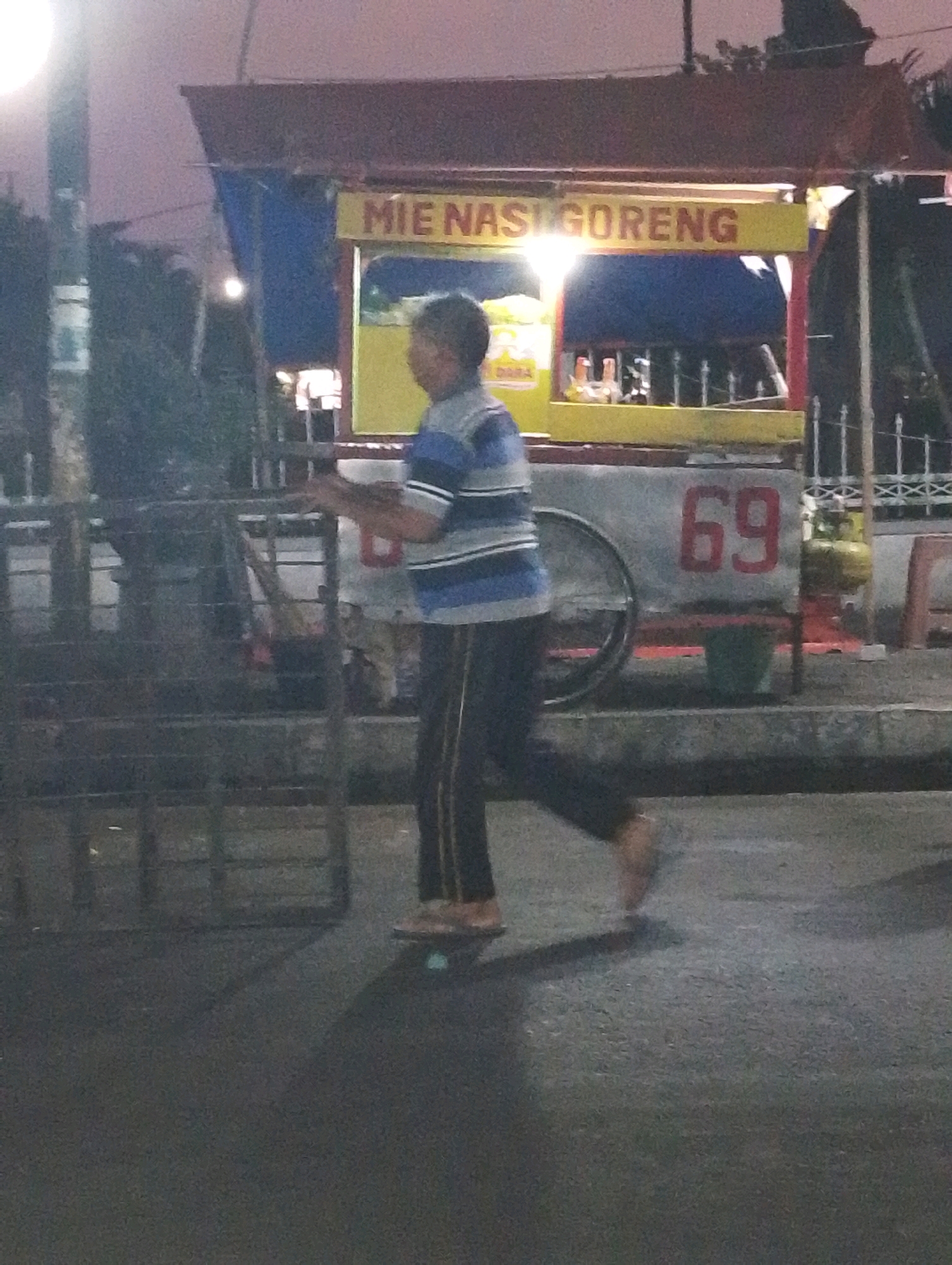 A full-body, side-view, motion-blurred medium shot captures a young person walking on a paved street at dusk in Bangil, Indonesia. The person, who appears to be male, is wearing a blue and white striped short-sleeved shirt, black pants with a white stripe down the side, and brown sandals. They are walking from left to right, with their right arm extended forward and their left arm bent behind them, suggesting they are in mid-stride.
Behind the person is a food cart that displays the text "MIE NASI GORENG" in red letters on a yellow sign. The cart is parked on the side of the road, and the area behind it appears to be a commercial or street vendor zone. The cart has a blue tarp covering part of it and a prominent red "69" painted on its side. A propane tank is visible next to the cart. The ground is paved, and there are metal barriers and a fence to the left and right of the cart. The background shows trees and structures, with a hazy, dusky sky overhead. The lighting suggests it is either early morning or late evening.