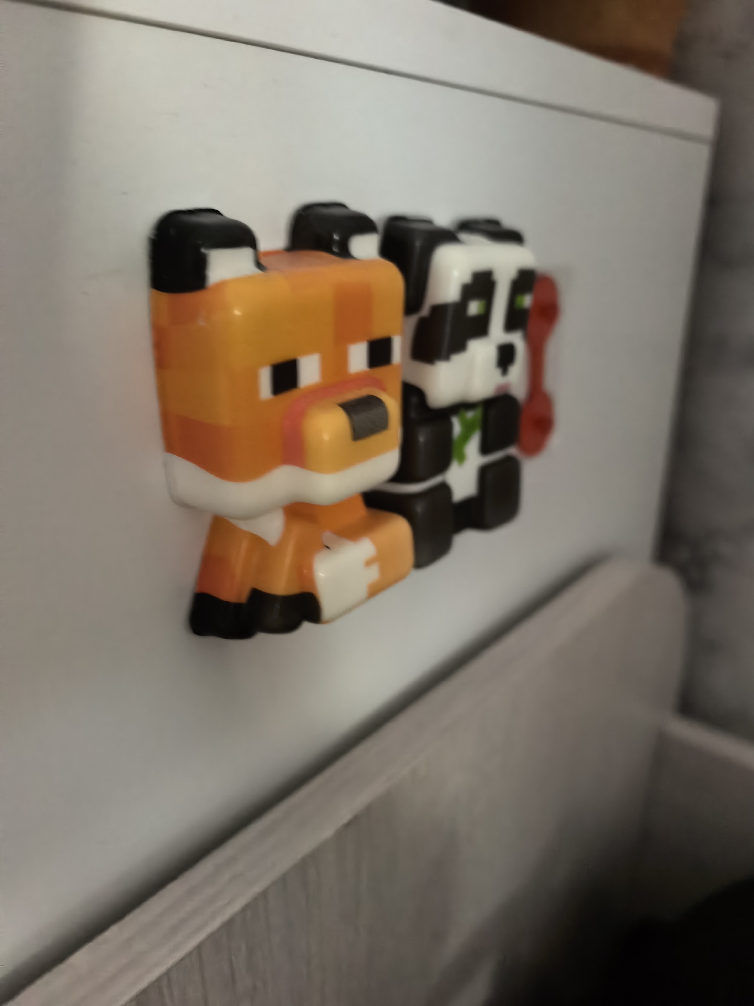A close-up shot shows three Minecraft-style toy figures attached to a white surface. From left to right, the figures are a fox, a panda, and an unidentified character with red coloring. The figures appear to be made of plastic and have pixelated designs characteristic of Minecraft. The surface they are attached to appears to be a cabinet or similar piece of furniture. The background is out of focus, but shows hints of a wall and possibly more furniture. The location is not discernible from the image. There is no evidence indicating that the photo was taken in Sterlitamak, Russia.