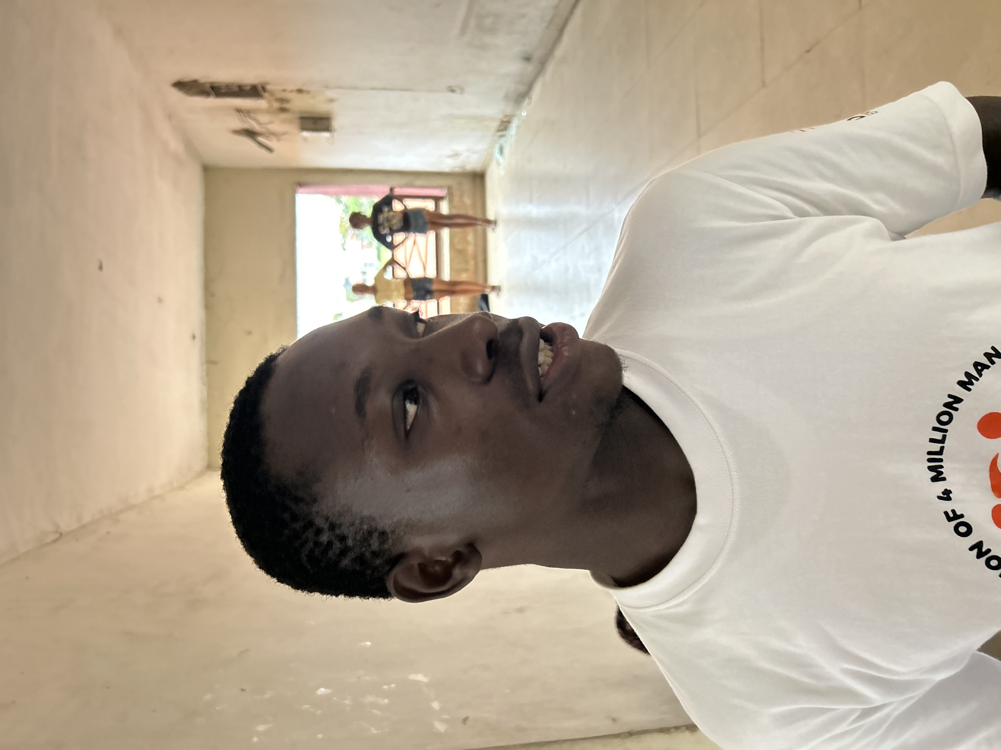 The image shows a young Black man in the foreground, looking to his left. He is wearing a white t-shirt with black text that reads "OF 4 MILLION MAN" and some orange graphics. The t-shirt has a slightly wrinkled texture. The man has dark skin, short black hair, and a faint beard. His mouth is slightly open, revealing his teeth.
In the background, there is a hallway with concrete floors and walls. The walls appear to be unpainted or have peeling paint, showing some exposed concrete. There are some wires and electrical boxes visible on the ceiling of the hallway.
Further down the hallway, through an opening, three other people are visible. Two of them appear to be young women, wearing t-shirts and shorts. They are standing and facing away from the camera. The third person is less distinct but also appears to be standing. The opening leads to an outdoor area with greenery visible, suggesting it might be daytime.
The overall setting appears to be an unfinished or under-construction building. The lighting suggests it is daytime. The man in the foreground seems to be the primary subject, and the context of the "4 MILLION MAN" on his shirt might relate to a specific event or organization. The image is taken in a portrait orientation.