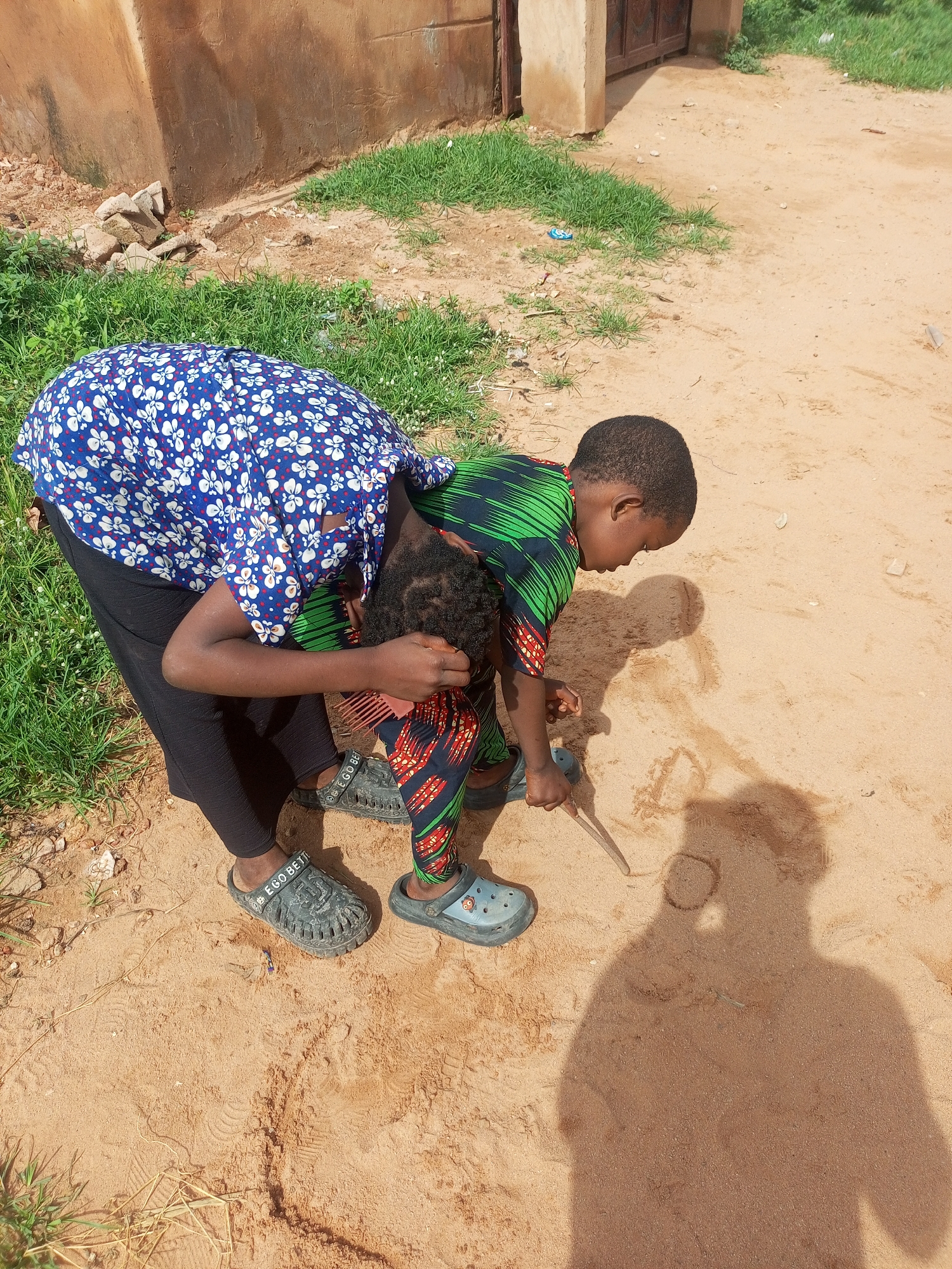 An overhead shot shows two children in Gombe, Nigeria, engaged in drawing in the sand. The child in the foreground, wearing a green and black patterned outfit with red accents, is leaning down, using a stick to etch designs into the sandy ground. Their shadow is cast across the sand, forming a large silhouette. Behind them, another child, dressed in a blue shirt with white floral patterns and black leggings, is also bent over, their face hidden from view. They appear to be assisting or observing the drawing. The ground is a mix of sand and sparse green grass, with a weathered brown wall and a metal gate visible in the background. The scene suggests a warm, outdoor environment, typical of many Nigerian cities.