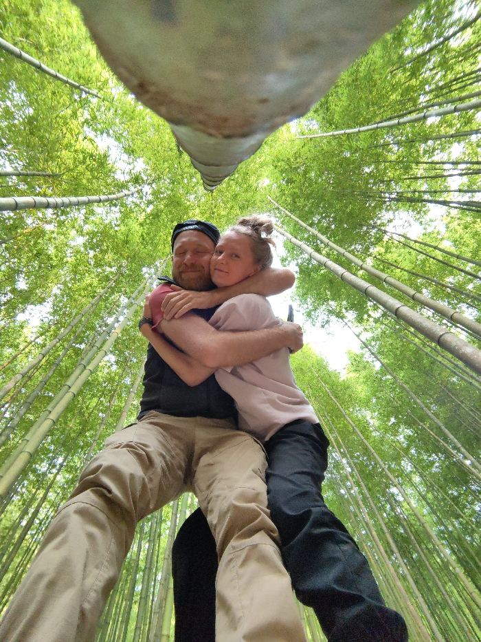 A low-angle, wide-shot photograph taken in a bamboo forest in Mù Cang Chải, Vietnam, depicts two individuals, a male and a female, embracing. The camera is positioned on the ground, looking upward, with a large, out-of-focus object (likely a bamboo stalk) occupying the top center of the frame. The male, on the left, has a beard, wears a dark backward baseball cap, a dark short-sleeved shirt with a red undershirt or sleeve visible, and tan cargo pants. His eyes are closed or squinting, and he displays a slight smile. The female, on the right, has light hair pulled into a bun, wears a light pink t-shirt and dark pants. She looks directly at the camera with a subtle smile. Both individuals have their arms wrapped around each other in a close hug. Surrounding them, numerous thin bamboo stalks extend vertically toward a bright, partially visible sky, with dense green foliage forming the canopy.