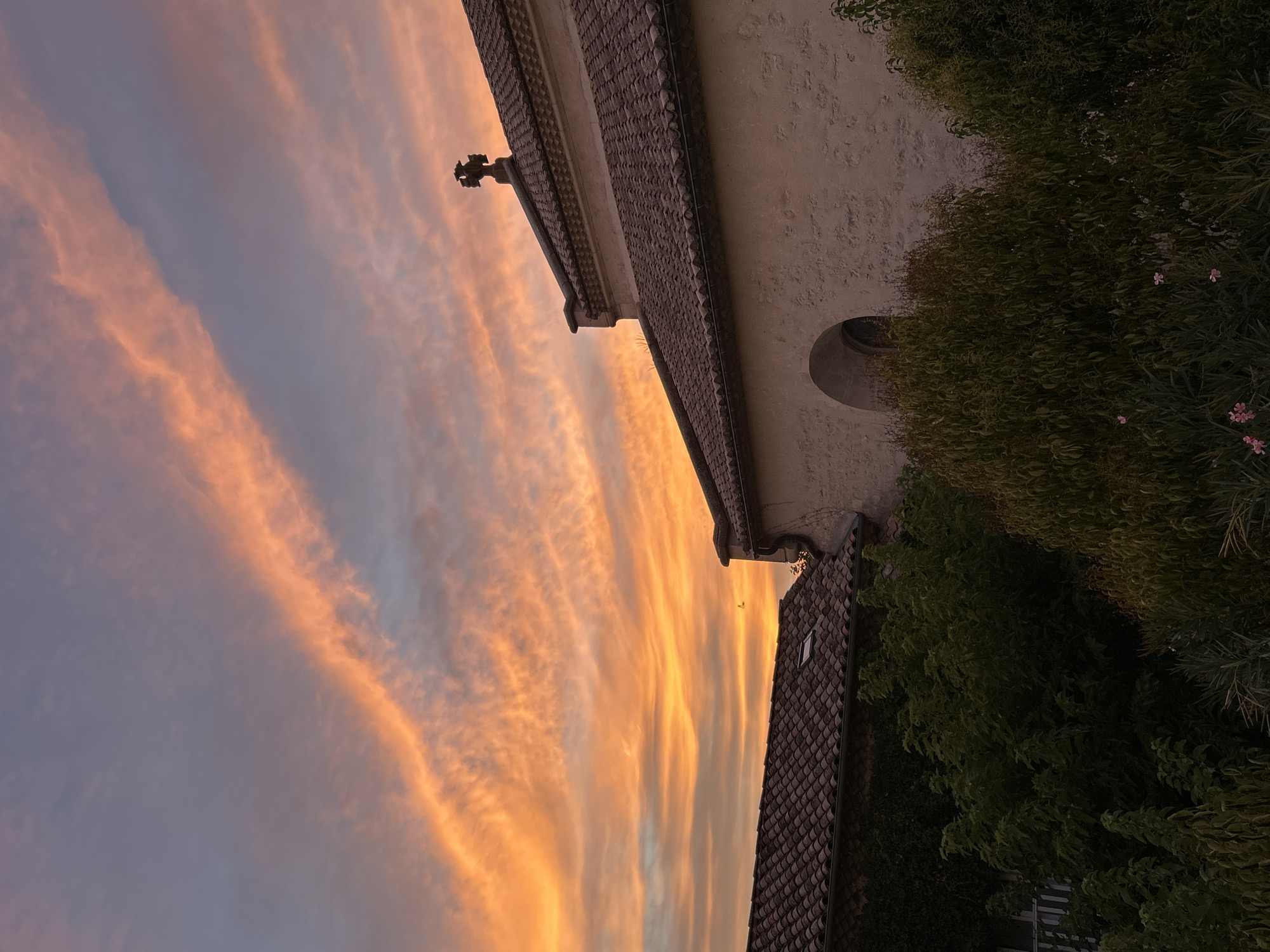The image captures a sunset in Saint-Marcel-lès-Sauzet, France, with the sky dominated by vibrant orange and pink clouds stretching across a dusky blue expanse. In the foreground, the terracotta tiled roofs of two buildings are visible, with one building featuring a textured stone facade and an arched window. A cross adorns the peak of the roof of the larger building. Lush green foliage, including trees and bushes, partially obscures the lower portions of the buildings. A small bird is visible in flight against the colorful sky. The scene conveys a sense of twilight calm over a village setting.