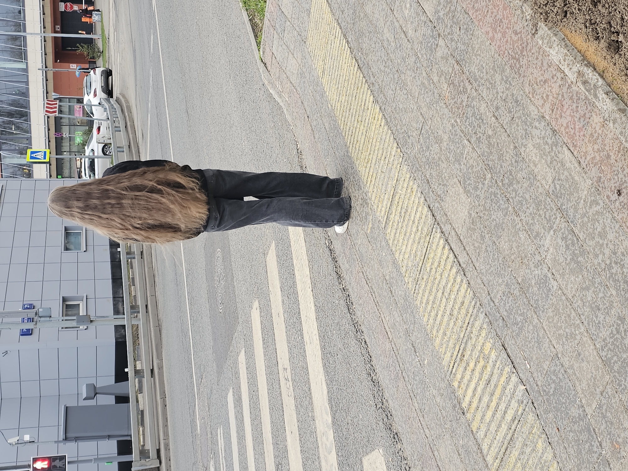A full-length shot reveals a person with long, light brown hair standing on a crosswalk in Khimki, Russia. They are wearing a black jacket and black jeans with white shoes. The crosswalk is marked with white stripes and is adjacent to a sidewalk with yellow textured stripes. In the background, there is a modern building with a gray facade. The street is visible with several parked cars and traffic signs. The sky is not visible.