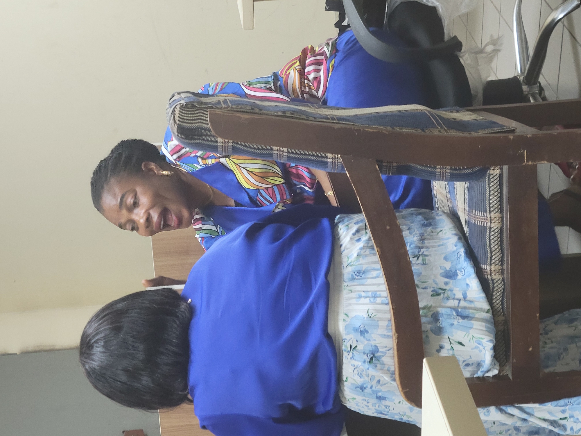 Two women are inside, near a chair. One woman is seated. The other woman is standing next to her. The seated woman is wearing a blue dress and has short, dark hair. The standing woman is wearing a patterned top and blue bottoms. The location is likely Abuja, Nigeria.