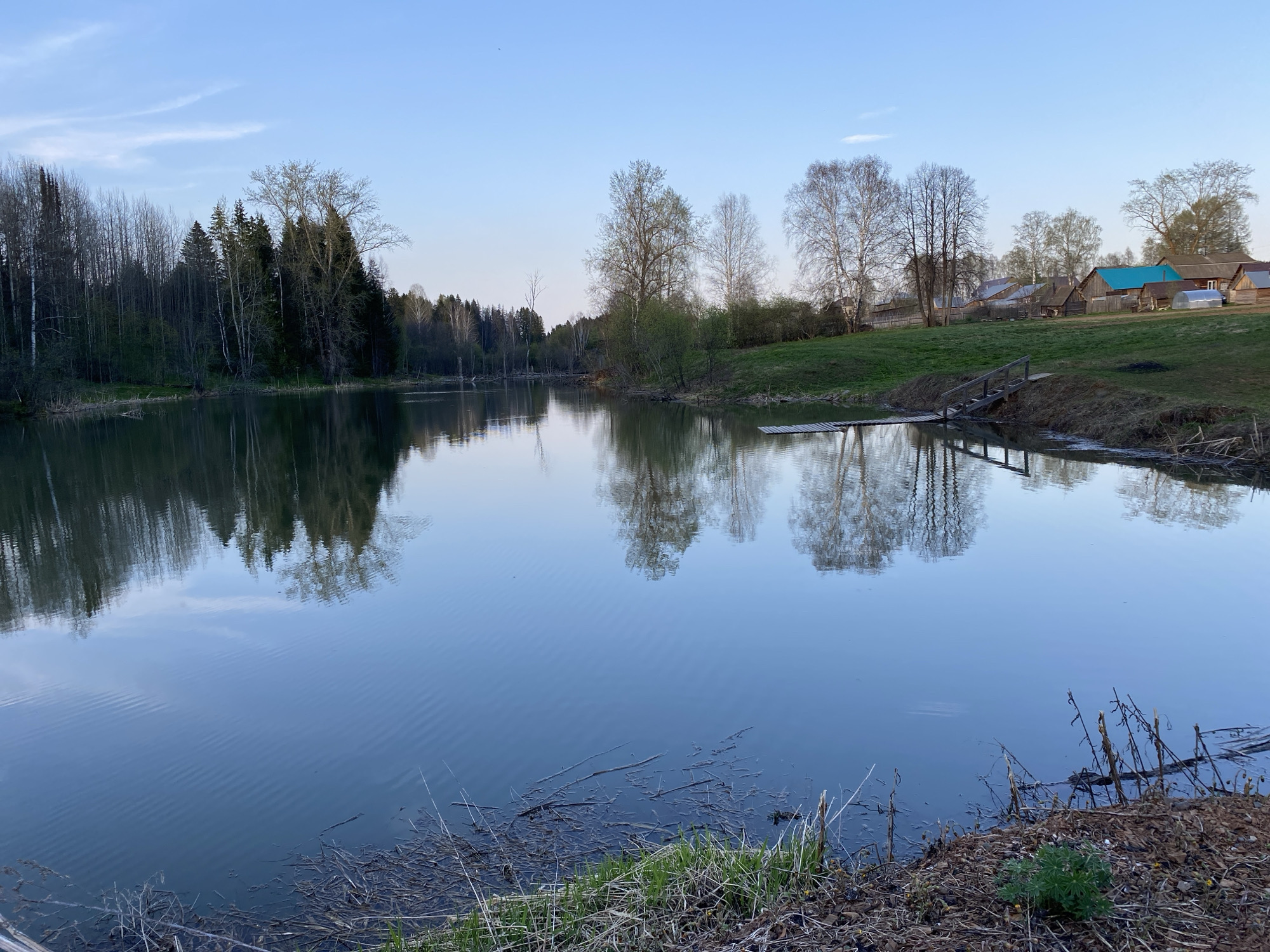 The image depicts a tranquil body of water, possibly a pond or lake, with reflections of trees on its surface. The body of water is surrounded by vegetation and trees. In the background, a small village or settlement is visible with houses featuring various roof colors. The scene appears to be located in a rural area, given the natural surroundings. The image does not provide enough information to confirm whether this image is from the city of Баклуши, Russia.