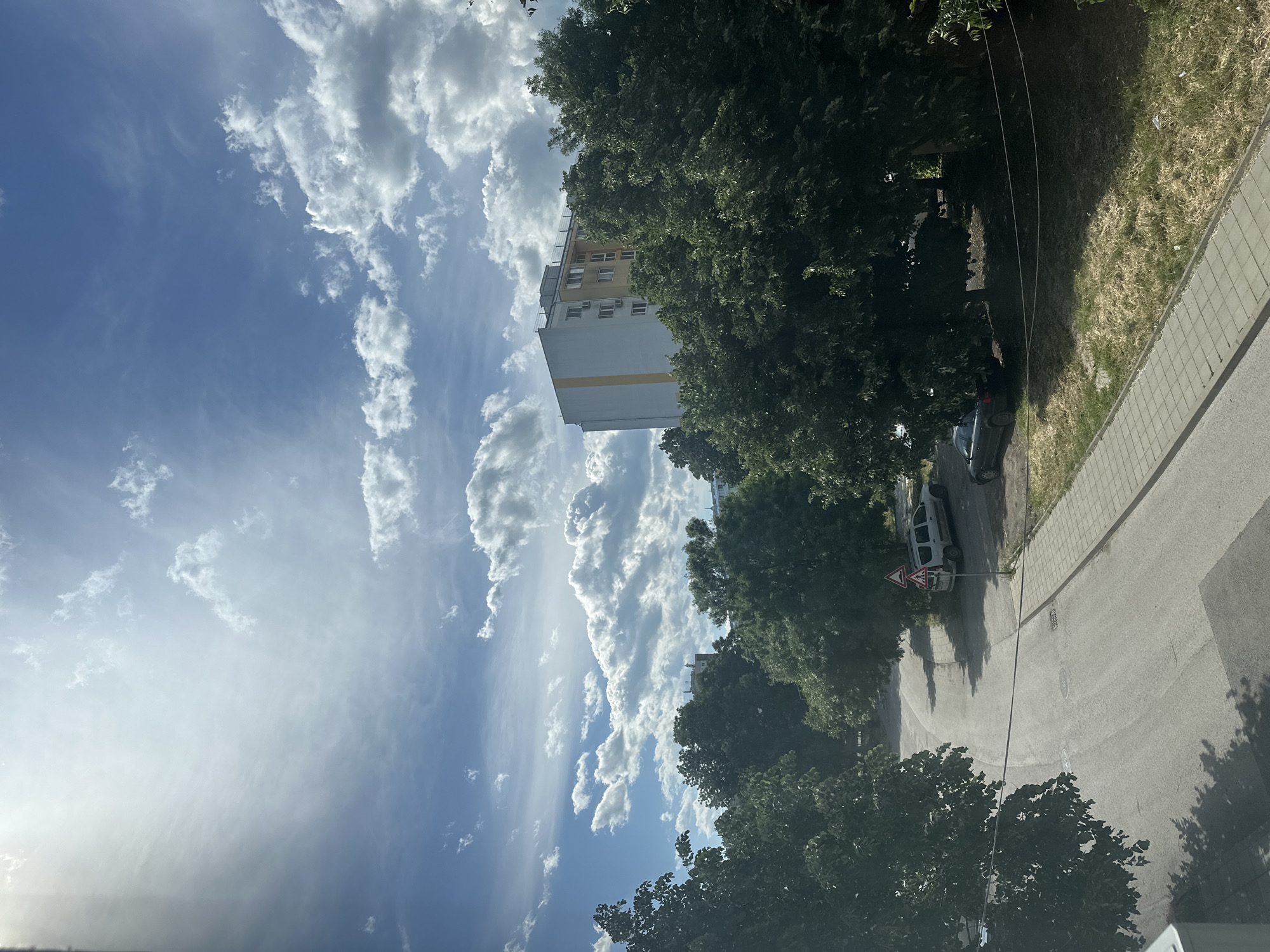 The image presents a street scene in the city of Unknown, Unknown. In the foreground, there is a paved road running in an upward diagonal direction. Green trees line the roadside, casting shadows on the pavement. Parked cars are visible alongside the road, next to a street sign. In the distance, there is a tall building, partially obscured by trees, against a backdrop of a blue sky filled with numerous white, fluffy clouds.