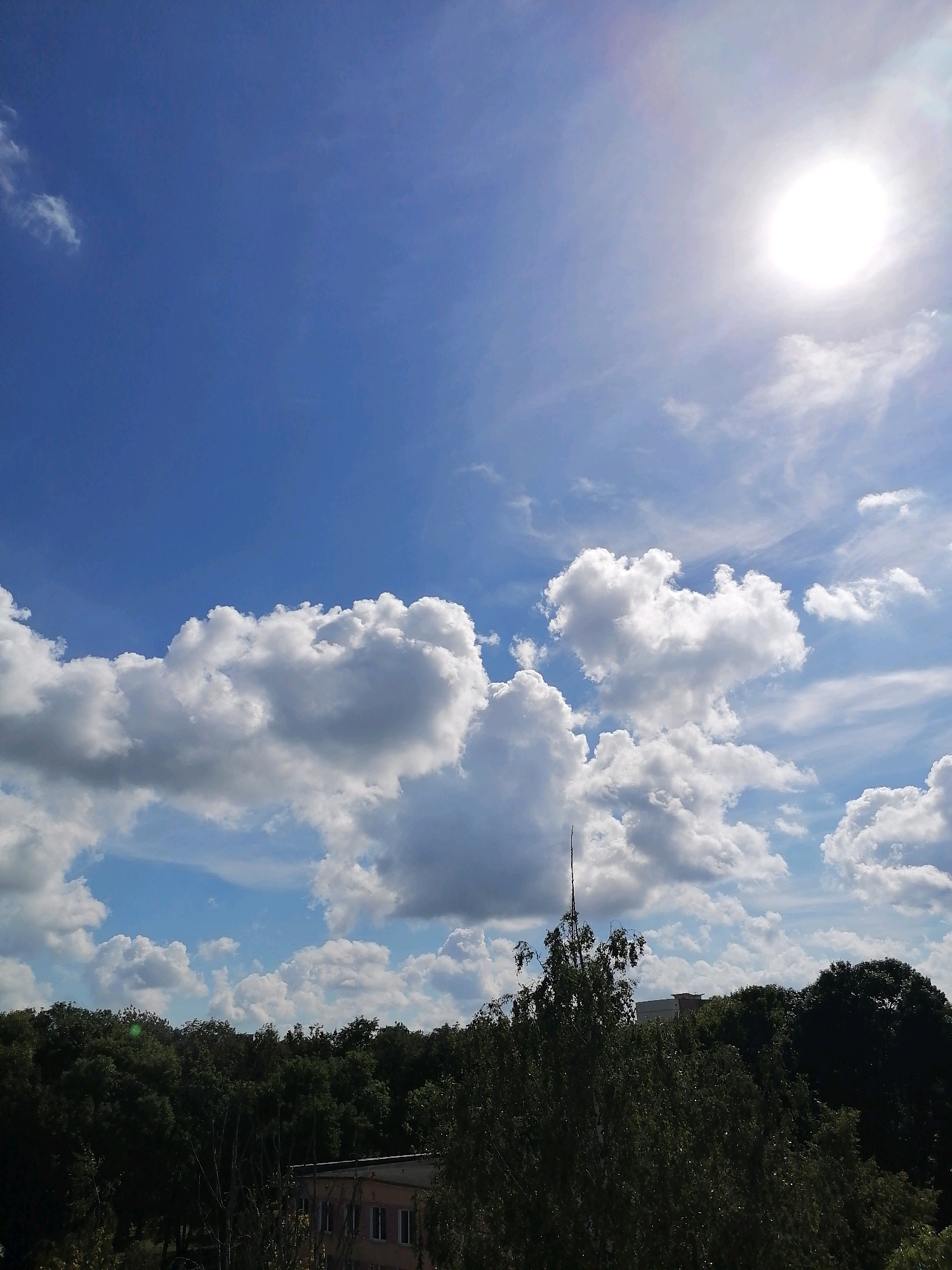 The image captures a bright, sunny day with a clear blue sky filled with fluffy white cumulus clouds. The sun is visible in the upper right corner, casting a bright light and creating a slight lens flare. In the distance, a dense line of green trees forms the horizon, with hints of buildings peeking through the foliage, suggesting an urban or suburban setting. The trees are dark green and appear to be a mix of deciduous and coniferous species. The lower portion of the image shows a building with a light-colored facade and several windows. The overall scene conveys a pleasant, summer day. The specific location of Hrodna, Belarus, cannot be determined from visual cues alone.