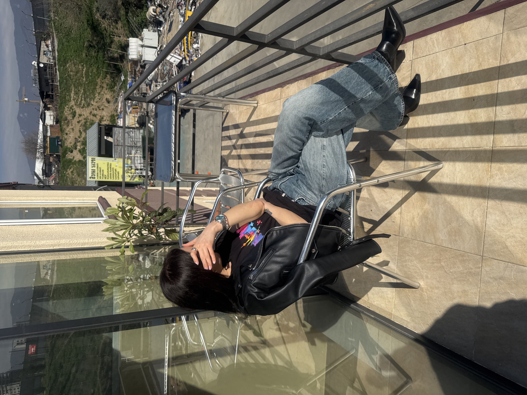 The image shows a woman lying in a metal chair on a balcony. She is wearing blue jeans, a black t-shirt with a colorful design, a black leather jacket, and black pointy-toe boots. Her face is partially obscured by her hand.
The balcony has a metal railing and tiled flooring with shadows cast by the railing. The backdrop includes an urban environment with various buildings and structures, suggesting a city. In the background, there are visible mountains and sky. There is greenery visible, possibly from a garden.
Based on the architectural style and overall environment, the location is possibly in Tbilisi, Georgia.
