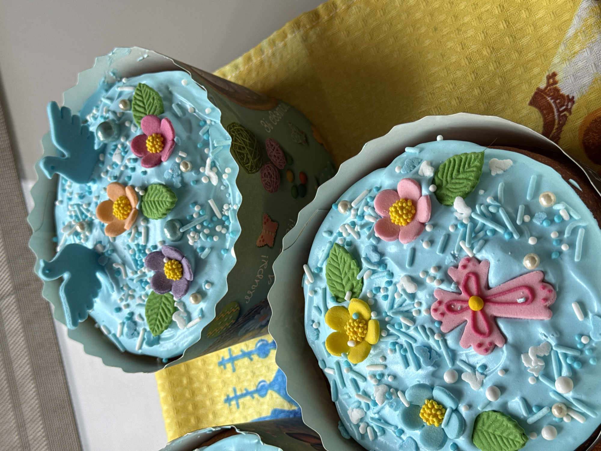 The image shows two decorated Easter cakes (Kuliches) on a yellow patterned cloth. The cakes are topped with light blue frosting, decorated with sugar flowers, green leaves, and sprinkles. One cake has a blue dove on it. The cakes are in paper wrappers, with the wrapper of the cake on the left decorated with images of colorful eggs and the text "Zi Sviatomi!" The image suggests a festive Easter celebration, likely in a home setting. Given the cake decorations and the phrase "Zi Sviatomi!", which is a Ukrainian phrase for "Happy Holidays!", the cakes were likely made in Kyiv, Ukraine.
