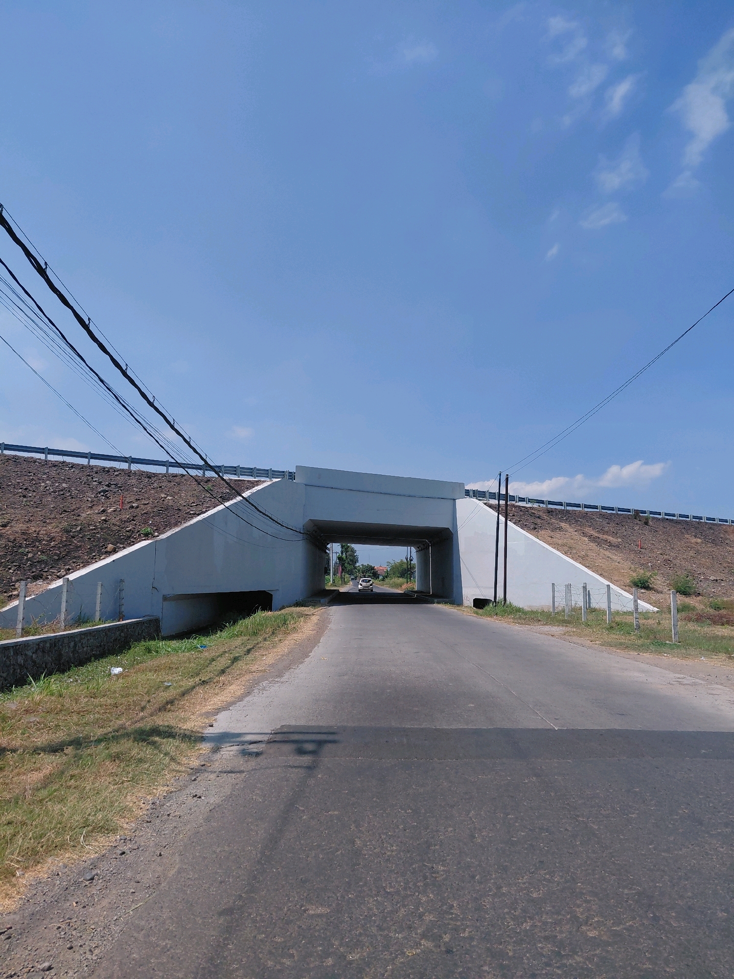 A wide-angle, eye-level shot captures an underpass on a clear, sunny day in Sumberan, Indonesia. The underpass, constructed of white concrete, is centered in the frame, allowing a road to pass beneath it. The road leading to the underpass is paved and appears to be in good condition, with a clear lane marking.
On either side of the road leading to the underpass, the terrain slopes upward, covered with dry grass and loose rocks. A metal guardrail runs along the top of these slopes. Several dark power lines are strung across the sky, suspended by poles on the right side of the frame. In the distance, through the underpass, a white car is visible, suggesting ongoing traffic.
The sky is a vibrant blue with a few scattered white clouds. The overall impression is one of a functional piece of infrastructure in a rural or semi-rural setting, bathed in bright daylight. The scene appears to be free of people.