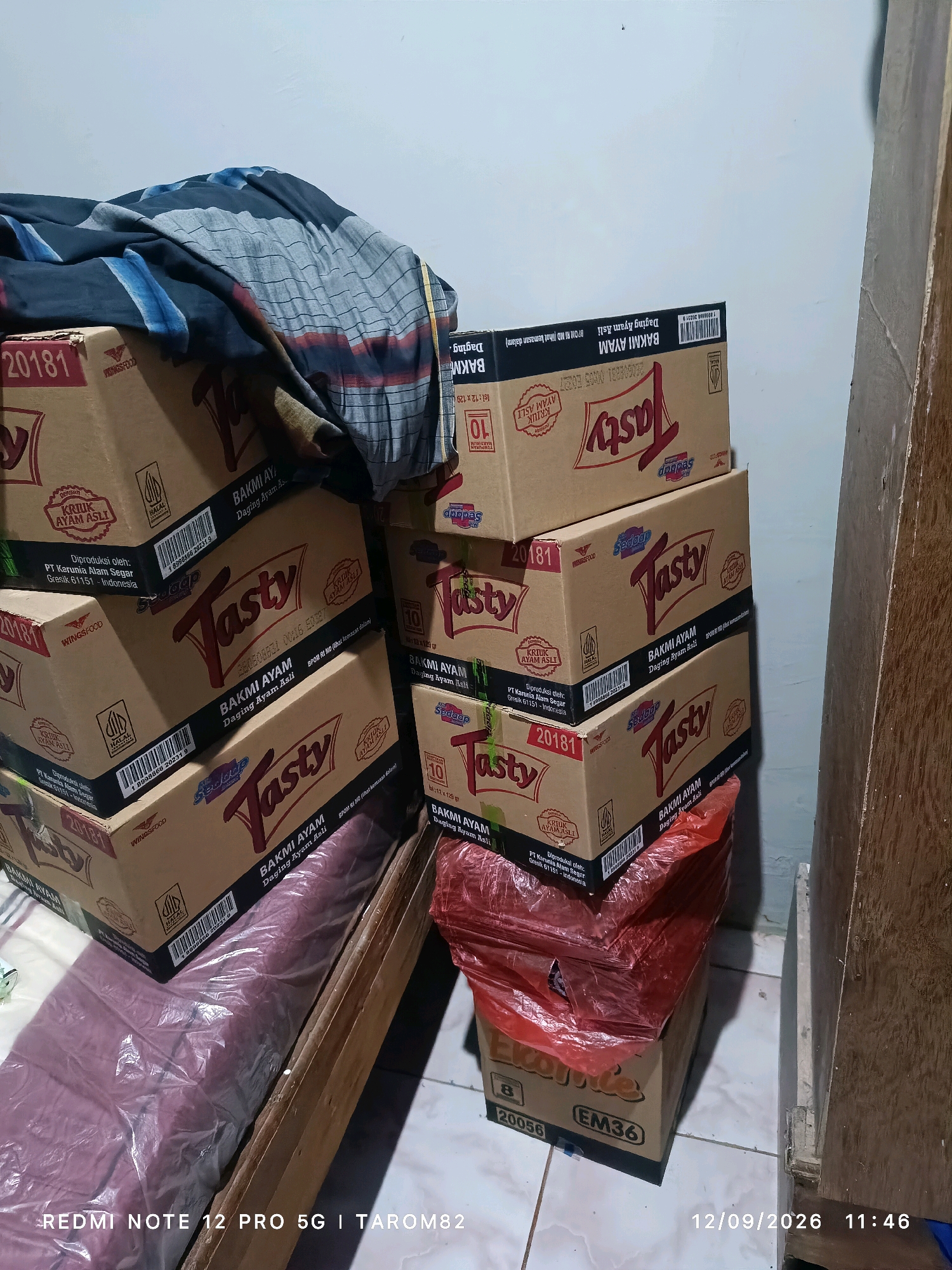 The image shows a stack of boxed instant noodles, branded "Tasty" and "Ekoje," with some text in Indonesian visible. The boxes are mostly brown cardboard with red and white lettering. Some boxes are labeled "Bakmi Ayam Daging Ayam Asli" which translates to "Chicken Noodles Real Chicken Meat." One box has the number "20181" and another "20056." There is also a red plastic bag containing items that are not clearly discernible, placed on top of some of the boxes. A folded, patterned fabric, possibly a sarong, is draped over the top of the stacked boxes on the left side of the image. The background appears to be a wall and a wooden surface. The floor is tiled. The timestamp in the bottom right corner indicates the photo was taken on September 12, 2026, at 11:46 AM. The text in the bottom left corner reads "REDMI NOTE 12 PRO 5G | TAROM82", likely indicating the device used to take the photo and a username. The image is an indoor scene, likely a room in a residence or storage area, and there is no indication of the specific location within Rejoagung, Indonesia.