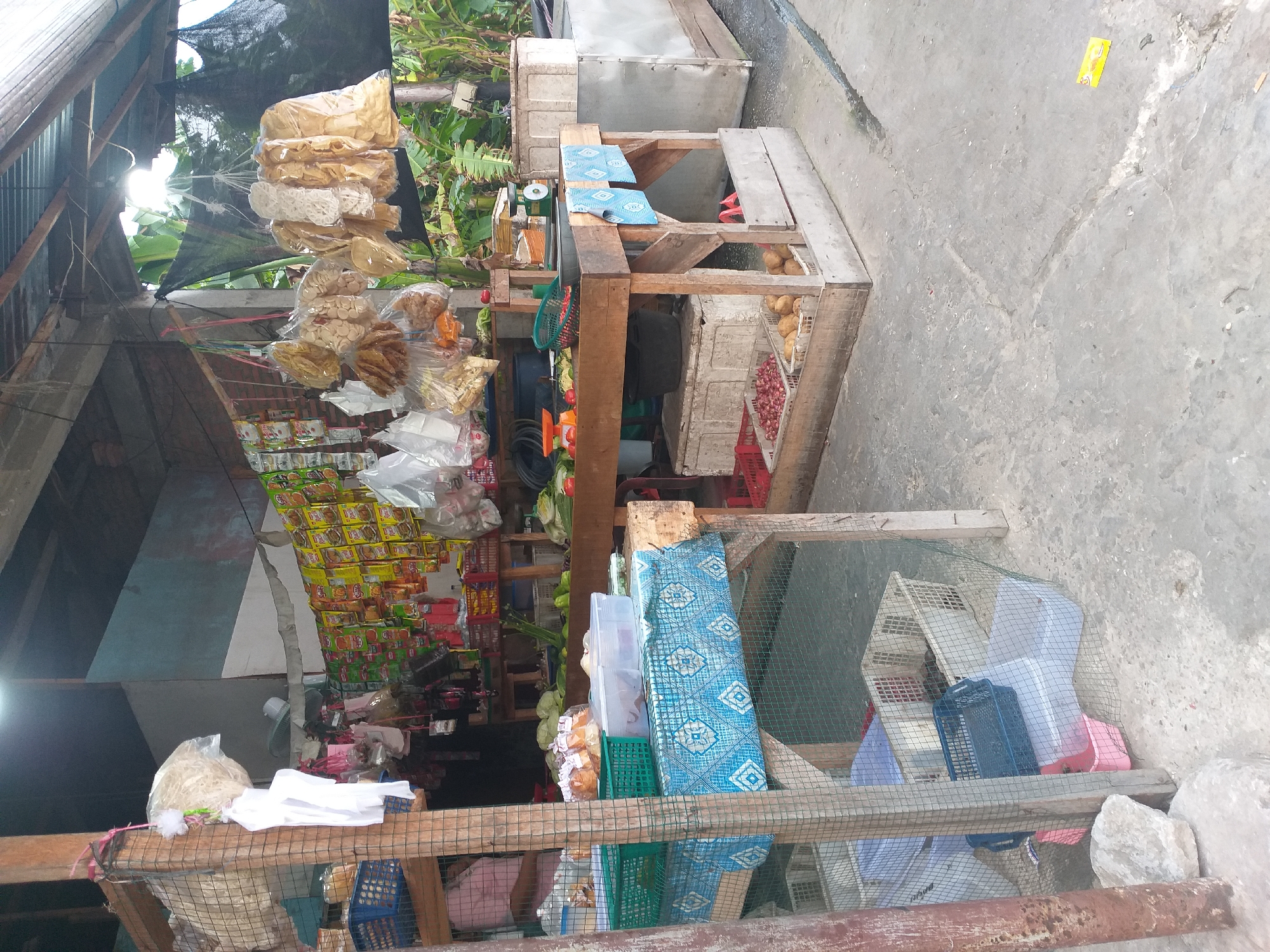 The image depicts an outdoor market stall in Air Jamban, Indonesia. The stall is constructed of wood and metal, with a corrugated metal roof providing shelter. Various items are for sale, including packaged snacks, vegetables, and what appear to be dried goods or crackers hanging from the ceiling. The stall is organized with shelves, baskets, and display tables. On one table, a scale and various vegetables are visible. Below the table, crates contain onions and potatoes. In the foreground, a display table is covered with a blue and white patterned cloth, with plastic containers and baskets on top. The ground is paved with concrete, and some debris is scattered around. The scene appears to be active, with goods displayed for sale in a typical market setting.