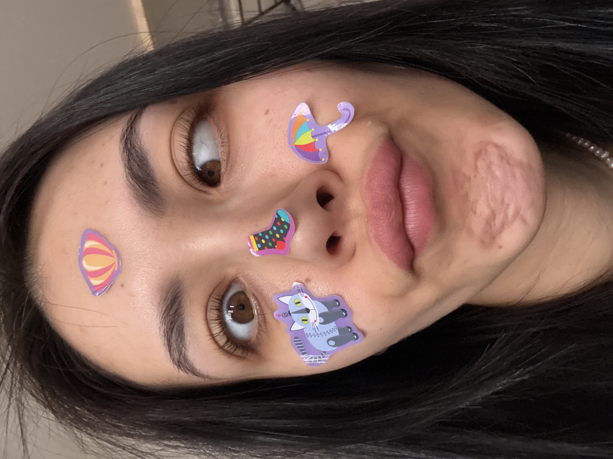 A close-up, slightly rotated shot features a woman's face adorned with various colorful stickers. A shell-shaped sticker is on her forehead, a purple umbrella-shaped sticker is near her nose, a polka-dotted sock is above her nose, and a cat robot sticker is on her cheek. The woman has dark hair, visible brown eyes, and light skin. There is a visible scar on her right cheek. The background is a solid neutral color. The location is unknown, not specified in the image.