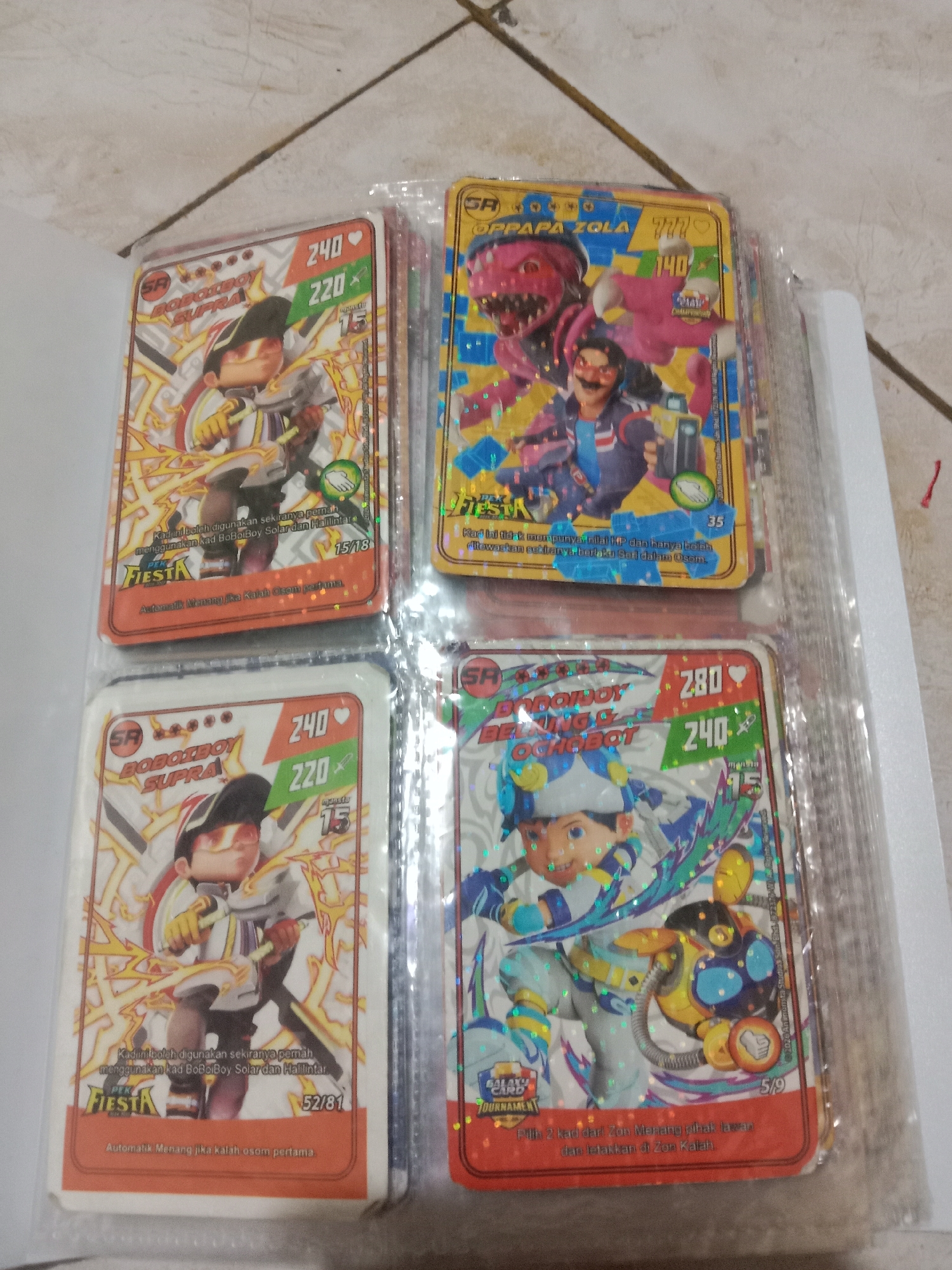 The image shows four trading cards inside a clear plastic sheet protector, which is lying on a tiled floor. The cards depict characters from the animated series Boboiboy. From top left, the cards are:
* **Top Left:** A card featuring Boboiboy Supra. It has a character portrait of Boboiboy in his Supra form, with numbers "240" and "220" and a "15" in a leaf icon. The card has "FIESTA" branding.
* **Top Right:** A card featuring Papa Zola and a pink dinosaur-like creature. It has "777" and "140" displayed prominently. This card also has "FIESTA" branding.
* **Bottom Left:** Another card of Boboiboy Supra, similar to the top left card, with numbers "240" and "220" and a "15" in a leaf icon. This card has "FIESTA" branding.
* **Bottom Right:** A card featuring Boboiboy Ying and Ochobot. It shows "280" and "240" displayed prominently, along with a "15" in a leaf icon and "GALAXY CARD TOURNAMENT" branding.
The floor appears to be tiled with a light-colored, possibly white or off-white, material. The lighting suggests an indoor environment. There are no people or discernible actions occurring in the image; it is a still photograph of the trading cards. The presence of the tiled floor and the general aesthetic are not specific enough to pinpoint a location within Madiun, Indonesia.