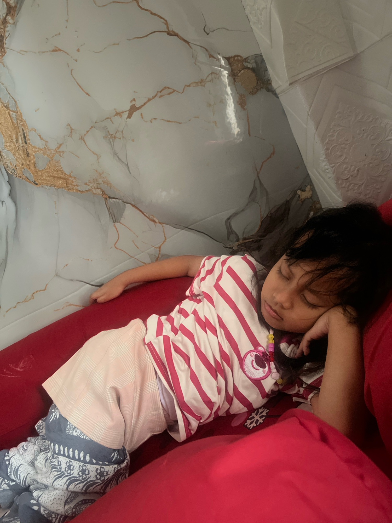 A young girl is asleep on a red couch. She is wearing a pink and white striped t-shirt and patterned shorts. Her head is resting on her hand, and she has a necklace with colorful beads around her neck. The background appears to be a wall with a marble-like pattern and some textured white panels. The scene is likely indoors, possibly in a residential setting in Kecamatan Pati, Indonesia, given the contextual information provided.