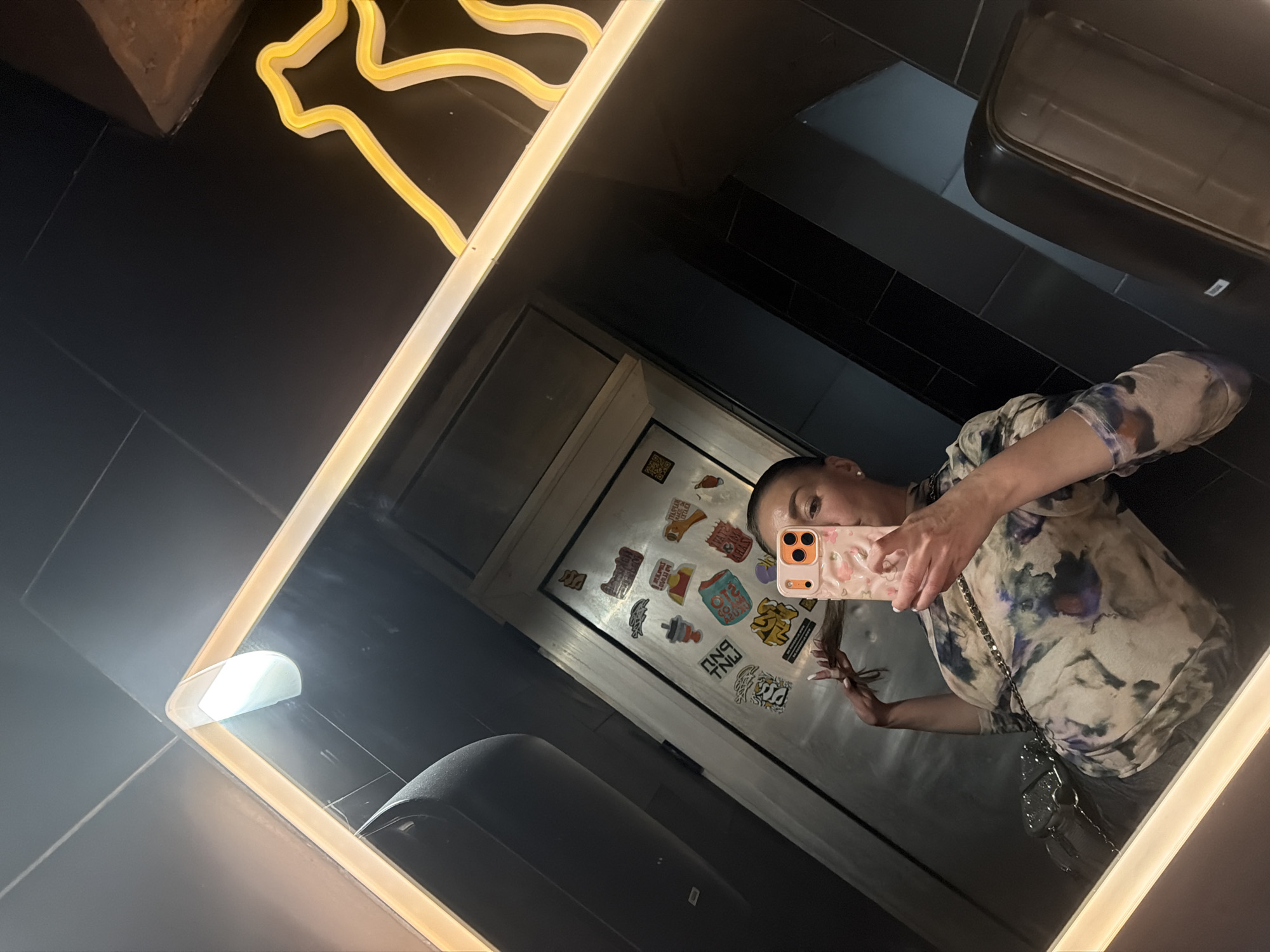 A woman takes a mirror selfie in a black-tiled bathroom. A hand dryer and a neon light adorn the walls. Stickers decorate the door, reflected in the mirror. The location is likely a commercial restroom in Bar, Montenegro, given the presence of the hand dryer and other standard fixtures.