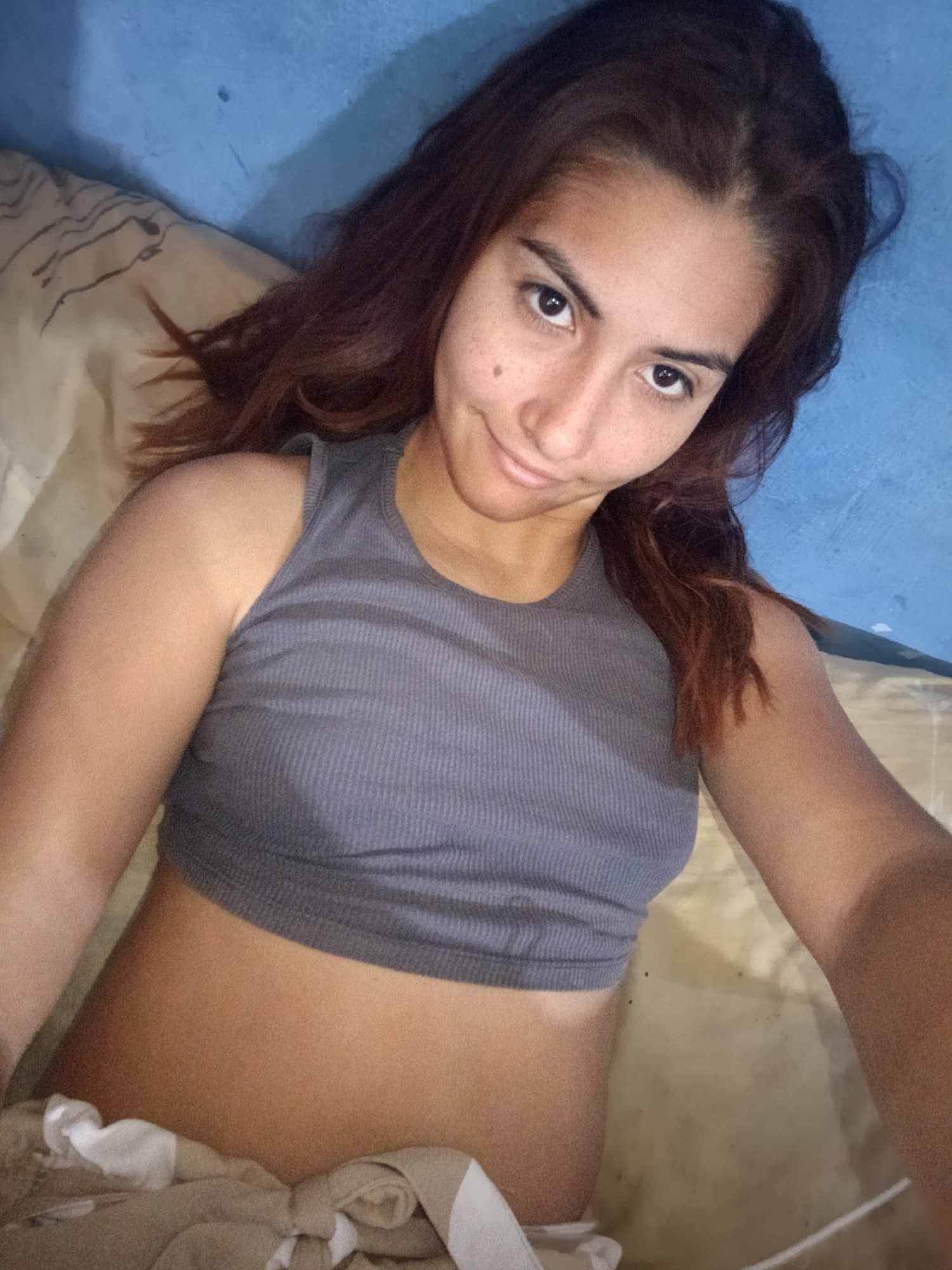 A woman is lying down, possibly in bed, with a blue wall in the background. She is wearing a grey crop top. A patterned pillow or blanket is visible. The location is unknown.
