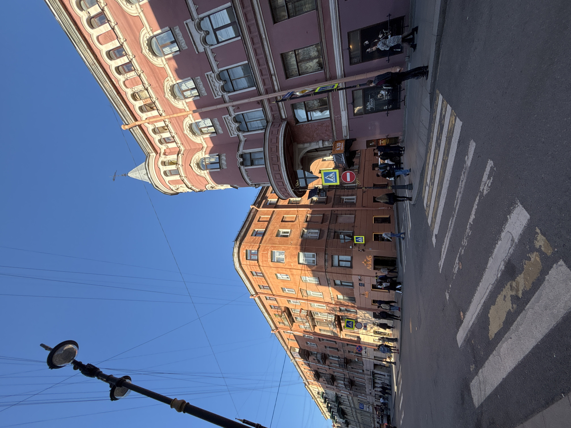 The image shows a street scene in Saint Petersburg, Russia. Two buildings, one pink and the other orange, flank the street. The pink building has ornate architectural details and arched windows, topped with a small conical roof. The orange building is more simple in design, with rectangular windows. People are visible walking on the sidewalks and crossing at a pedestrian crossing. Street signs and a lamppost are also visible. The sky is clear and blue.