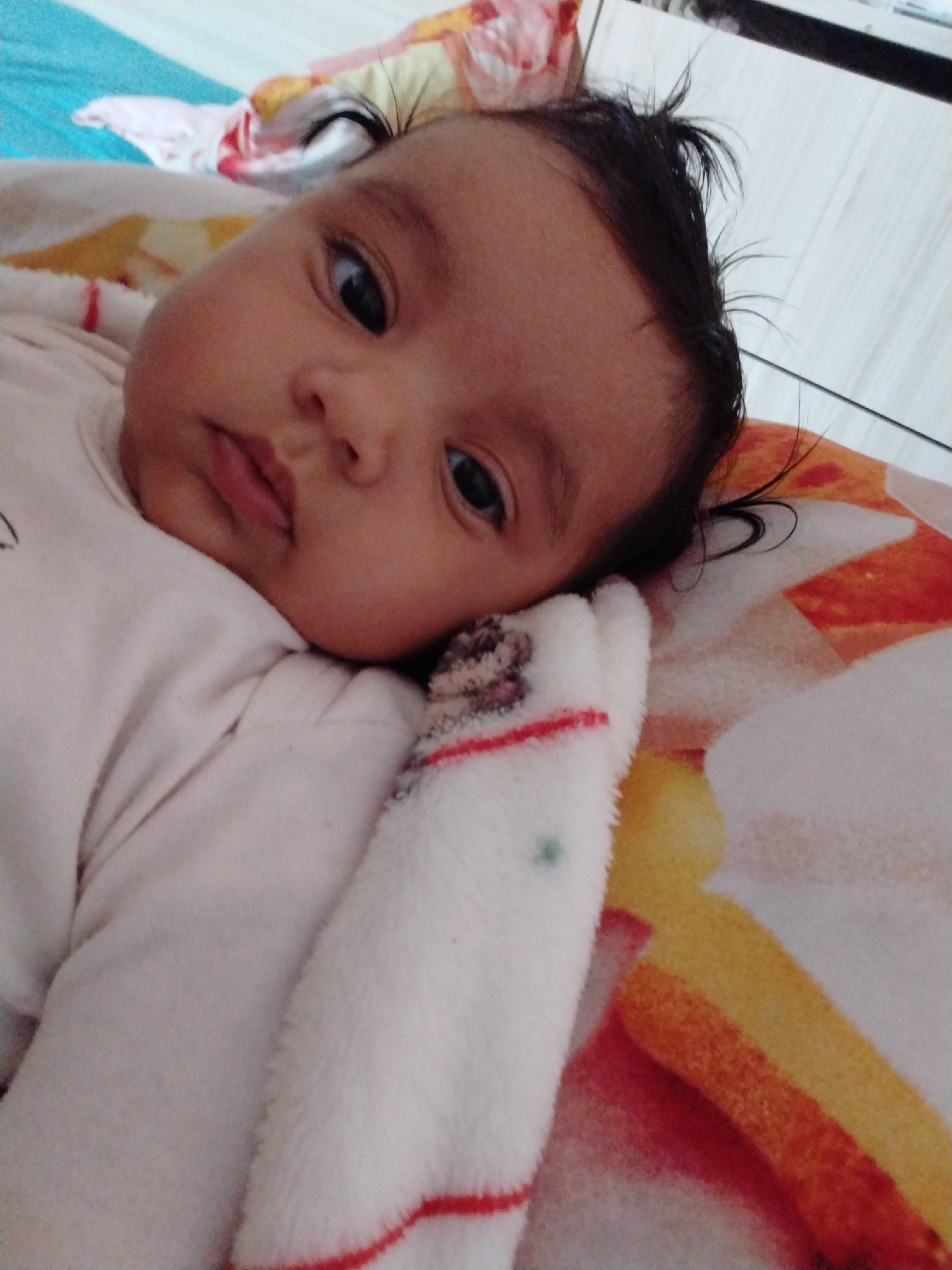 A baby lies on its side on a patterned blanket. The baby has dark hair and brown skin. It is wearing a light-colored onesie. The blanket has red, orange, and yellow stripes. Part of a white surface is visible in the upper right corner. Another blanket, blue, white, and pink, is visible in the top left. The location is Unknown, Unknown.