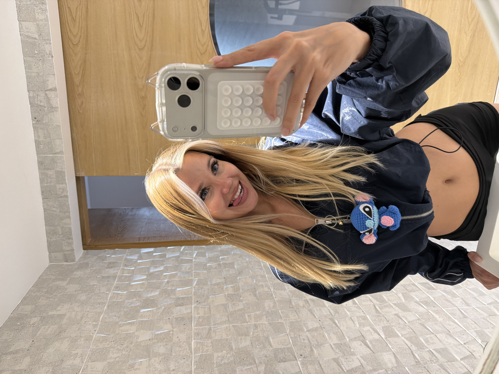 A young Caucasian woman with blonde hair and blue eyes takes a mirror selfie in a modern bathroom. She is wearing a dark blue cropped windbreaker with a Stitch keychain attached to the zipper and black shorts. Her stomach is exposed. She is sticking her tongue out at the camera. The bathroom features a tiled wall and a wooden door. The image was taken in Thep Krasatti, Thailand.