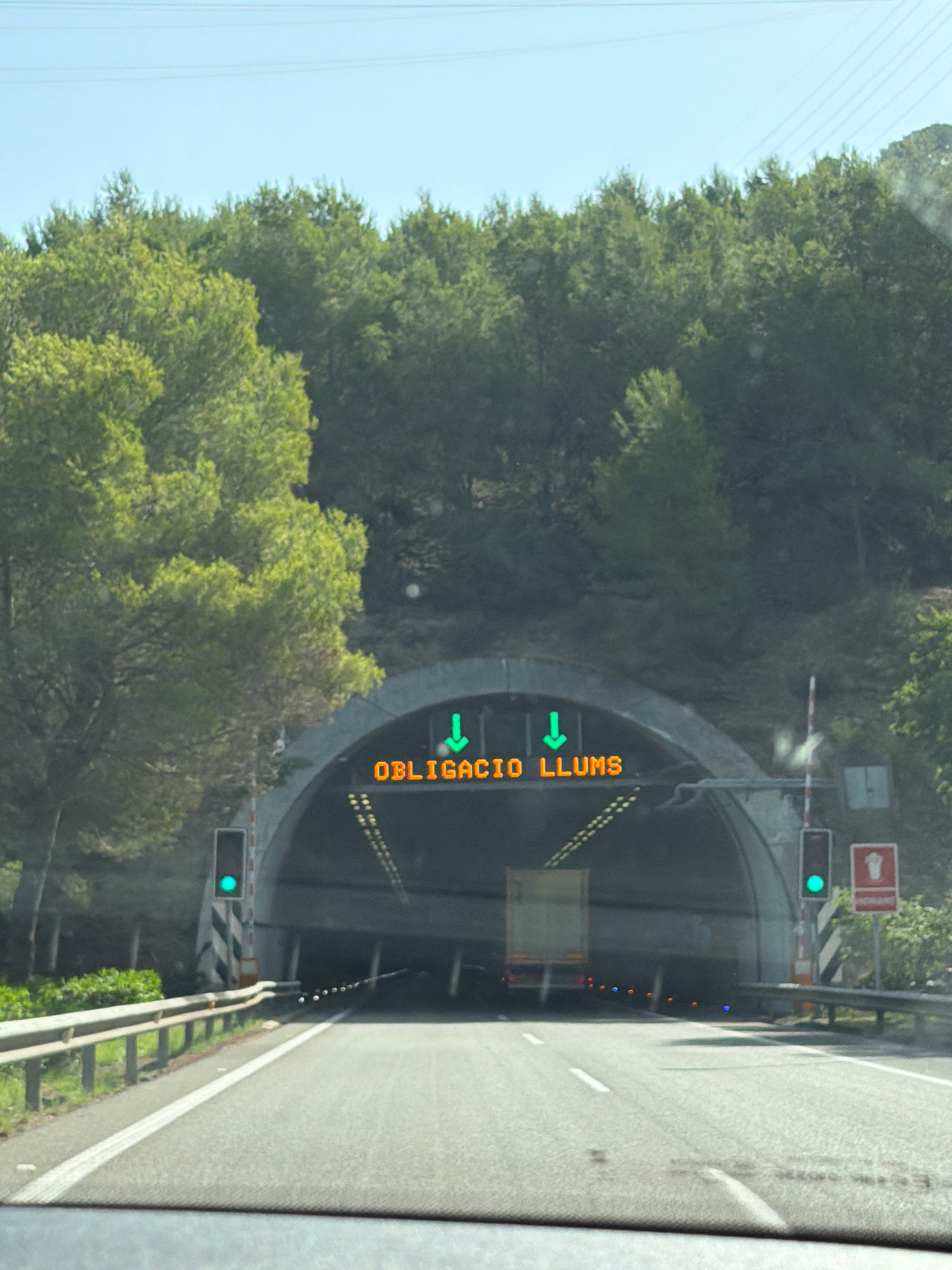 The image shows a view from inside a vehicle approaching a tunnel entrance on a sunny day. The road leading to the tunnel is paved with clear lane markings. On the left side of the road, there is a guardrail. Outside the tunnel, on the left, a traffic light is illuminated green. Above the tunnel entrance, a digital sign displays "OBLIGACIO LLUMS" in orange letters, with two green downward arrows. The tunnel is a large, arched structure. Inside the tunnel, a truck is visible, and there are other lights further within the tunnel. On the right side of the tunnel entrance, another traffic light is green, and a red and white sign with a pictogram is mounted on a pole. The background consists of a densely wooded hillside with green trees under a clear blue sky. Based on the language on the sign ("OBLIGACIO LLUMS" which is Catalan for "Obligation Lights"), this scene is likely on a road in Catalonia, Spain, potentially near Calafell, given the context provided.