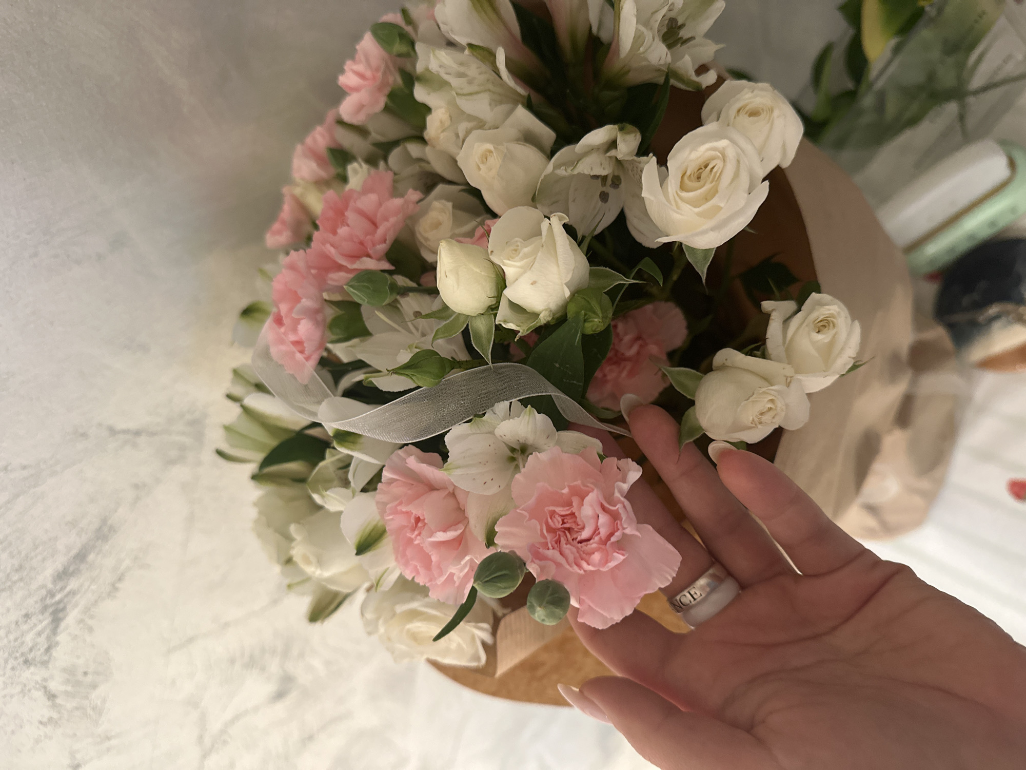 A close-up shot captures a bouquet of flowers, including white roses and pink carnations, wrapped in brown paper. A hand with polished nails and a silver ring touches one of the pink carnations. The background includes what appears to be a light-colored surface with a textured pattern, possibly a tablecloth or backdrop. The location is not identifiable based on visual cues in the image.