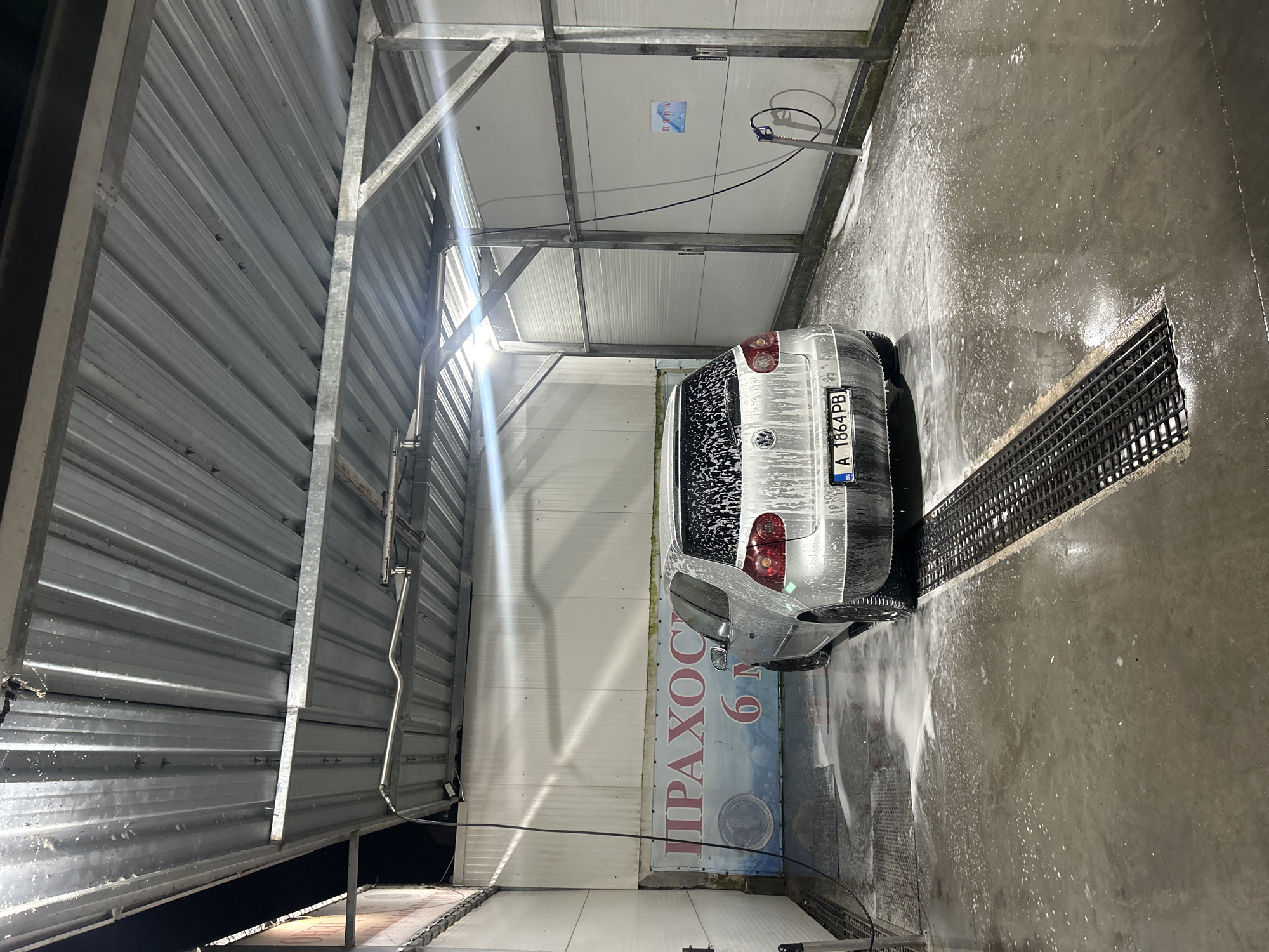 The image presents a silver Volkswagen Golf, likely a Mk5 model, positioned within a self-service car wash bay in Burgas, Bulgaria. The vehicle is viewed from the rear, heavily coated in white soap foam on its tailgate, rear window, and upper body panels, indicating it is actively being washed. The rear license plate, "A 1864 PB" with "BG" below, confirms Bulgarian registration and specifically designates Burgas by the 'A' regional code.
The car wash bay features a corrugated metal ceiling and white, vertically paneled walls, supported by an exposed metal framework of beams and trusses. Multiple bright artificial lights are affixed to the ceiling structure, illuminating the scene and creating lens flares. The concrete floor is wet with water and white soap suds, which collect and drain into long, dark metal grates embedded in the floor.
On the left side of the bay, a black high-pressure hose with a nozzle is visible, hanging from an overhead rail. A similar hose and blue-handled nozzle are suspended from a vertical metal pole on the right wall, adjacent to a wall-mounted control unit. A large banner on the back wall, partially obscured by the car, displays Cyrillic text including "ПРАХОС..." and "6 M", likely advertising a vacuum cleaner service. A smaller blue and red sign on the right wall reads "ПЯНА" (Foam) in Cyrillic. The scene is illuminated as if at night, and no individuals are present.