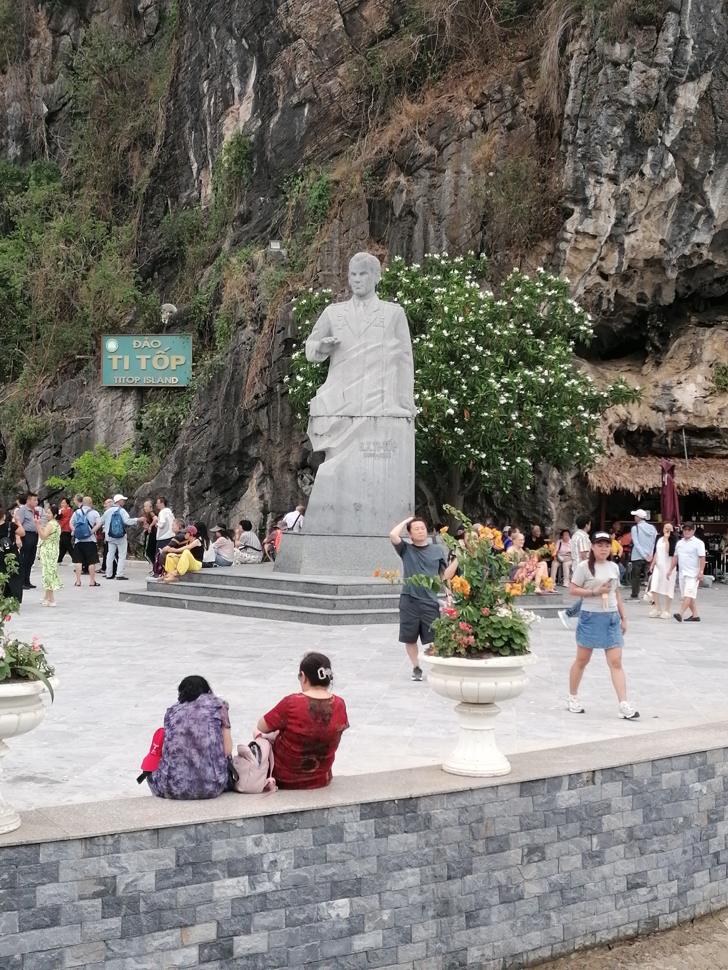 This image captures an outdoor plaza on Titop Island, located in Ha Long Ward, Vietnam, at the base of a dramatic, towering rock face. The central subject is a large, light-colored stone statue of a man in a suit, standing on a multi-tiered pedestal. Behind the statue, a vibrant tree with abundant white blossoms adds a touch of natural beauty against the rugged, weathered cliff, which is also partially covered in green and dry vegetation.
Numerous people are gathered in the plaza, suggesting a popular tourist destination. They are seen standing, walking, and sitting. Some individuals are resting on the steps leading up to the statue's base, while others are seated along a low, grey stone wall in the foreground. People are dressed casually in light clothing. In the immediate foreground, two women with their backs to the camera sit on a stone ledge next to a decorative white planter filled with colorful flowers. Another individual walks past the planter, and a man stands nearby, seemingly adjusting his hair.
A prominent green sign attached to the cliff face clearly displays "ĐẢO TI TỐP" in large Vietnamese script and "TITOP ISLAND" below it in English. The plaza floor is paved with grey tiles, and the low wall is constructed from grey rectangular stones. The lighting is bright but diffused, indicating either an overcast day or the softer light of late afternoon. The scene depicts a relaxed atmosphere typical of a sightseeing location.