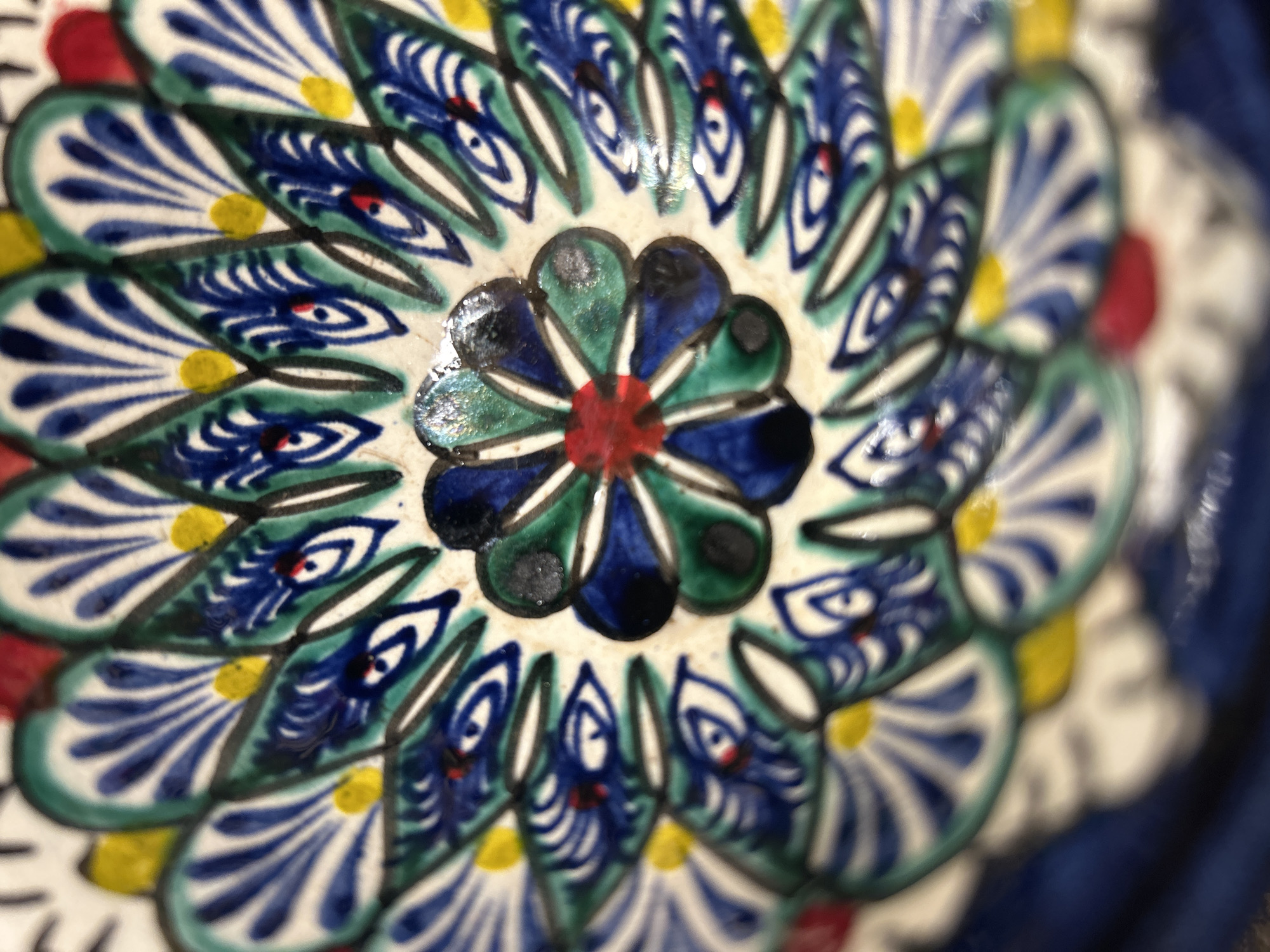 A close-up image of a ceramic plate with a complex floral pattern. The central design features a red circle at its core, surrounded by petals in shades of green and blue, outlined in black. A further layer of repeating, stylized floral elements in blue and white with green accents emanates outward. These elements include small red and yellow details. The background shows other parts of the design receding into blurry focus. The exact location is unknown.