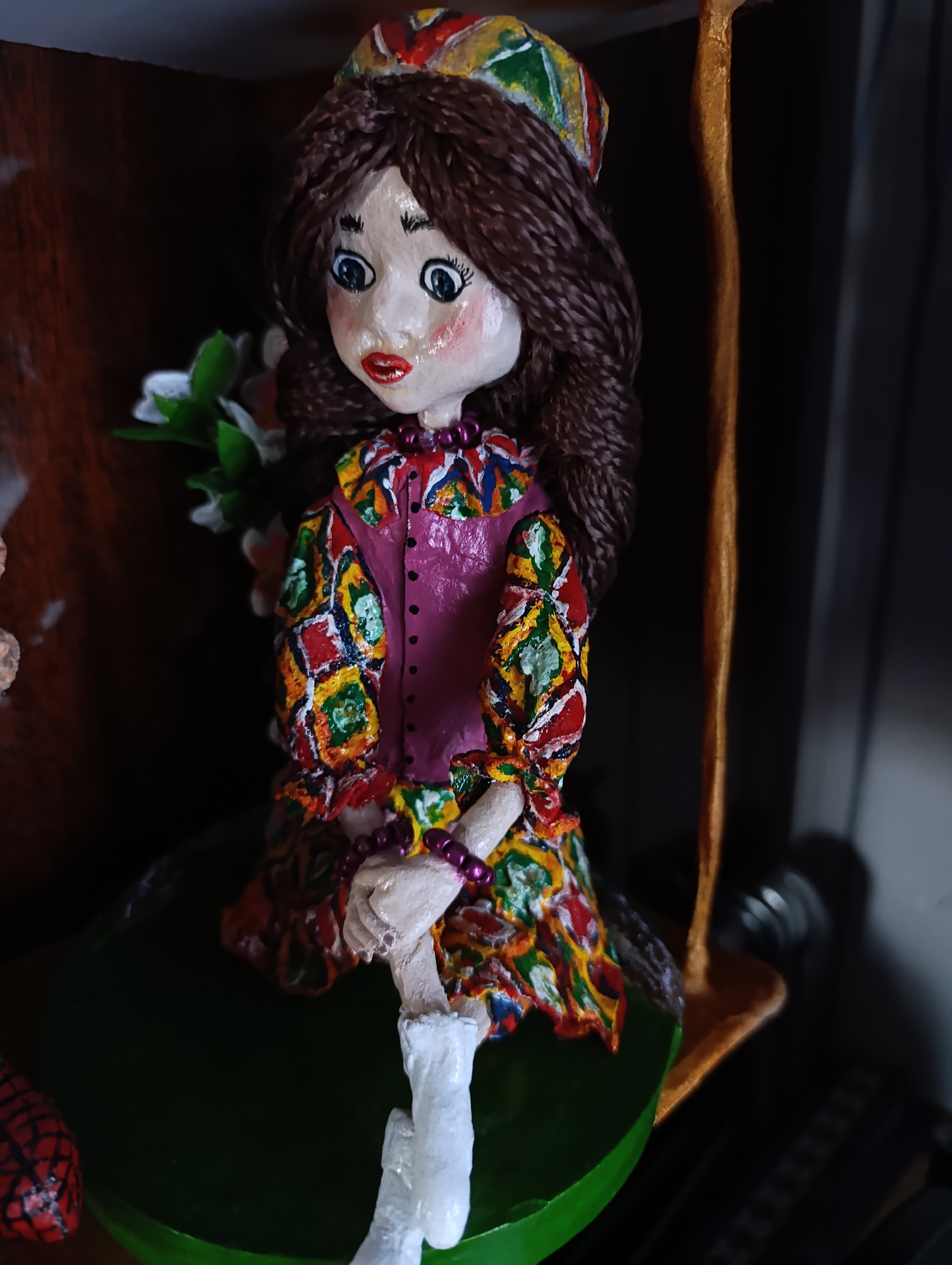 This media file features a close-up of a handmade doll, likely created from clay or similar material. The doll is positioned centrally, depicted as a young girl with brown braided hair, large blue eyes, and rosy cheeks. She wears a vibrant, colorful outfit with a patterned dress and a matching hat. Her dress features a detailed geometric design in red, yellow, green, and blue squares against a white background, with a pink vest over it. She also has purple beaded necklaces. The doll is seated on a green surface that appears to be a leaf or a piece of fabric, and is leaning against a golden support. In the background, there are hints of a wooden surface and some white flowers. The overall impression is that of a decorative folk art piece, possibly representing a traditional Uzbek costume from the Fergana Valley region, given the location context. The doll's facial expression is neutral, and the lighting suggests an indoor setting. No visible text is present in the image.
