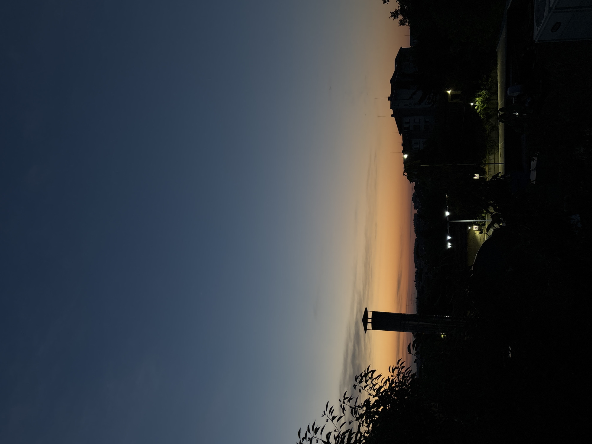 The image captures a dusky, twilight scene in the city of Pleven, Bulgaria. In the foreground, silhouetted foliage obscures the lower left portion of the frame. To the left of center, a tall, cylindrical chimney with a cap stands prominently, also in silhouette. In the middle ground, a street is visible, illuminated by several streetlights that cast pools of warm light. A few buildings, their structures indistinct in the low light, are visible beyond the street, with a larger building to the right. The sky transitions from a deep, dark blue at the top to a gradient of lighter blue, orange, and yellow hues near the horizon, indicating the sun has recently set. The overall impression is one of a quiet urban evening.